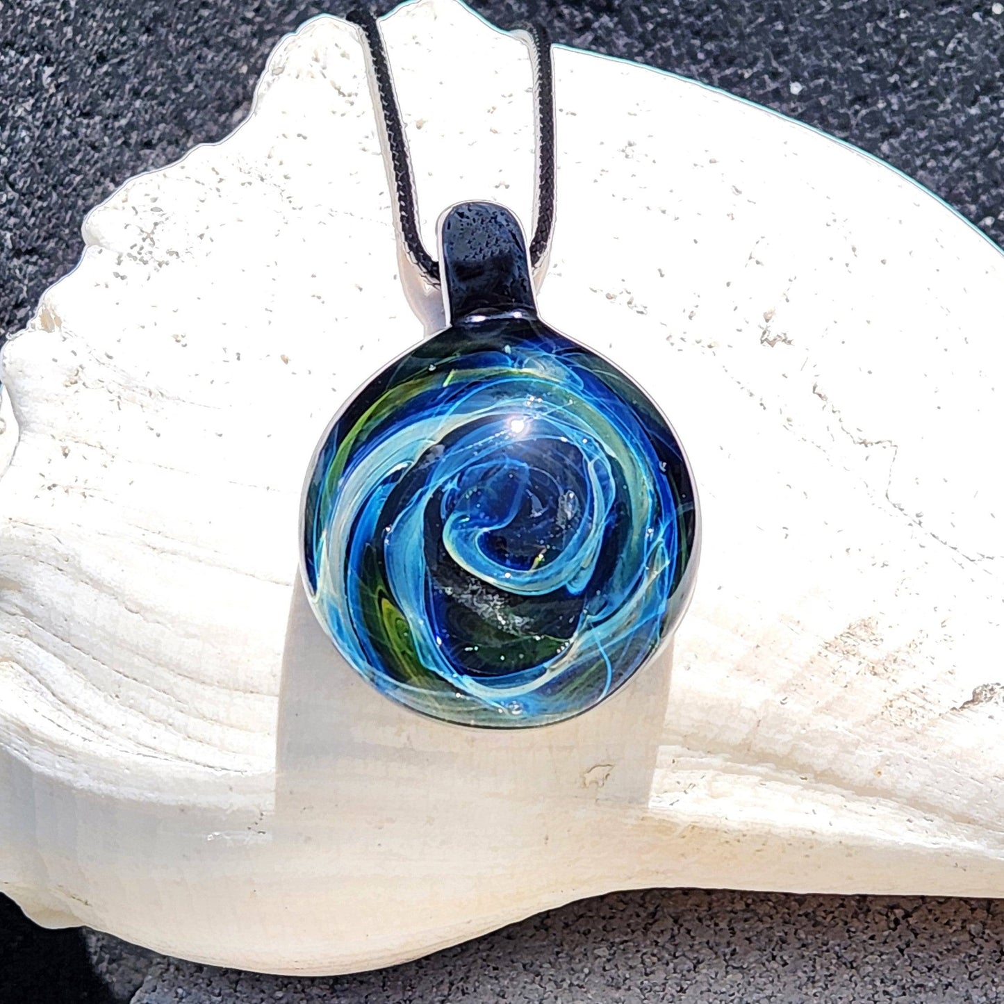 Heady Glass Pendant with swirling galaxy design in blue, green, and gold tones, handmade from borosilicate glass, displayed on a white seashell.