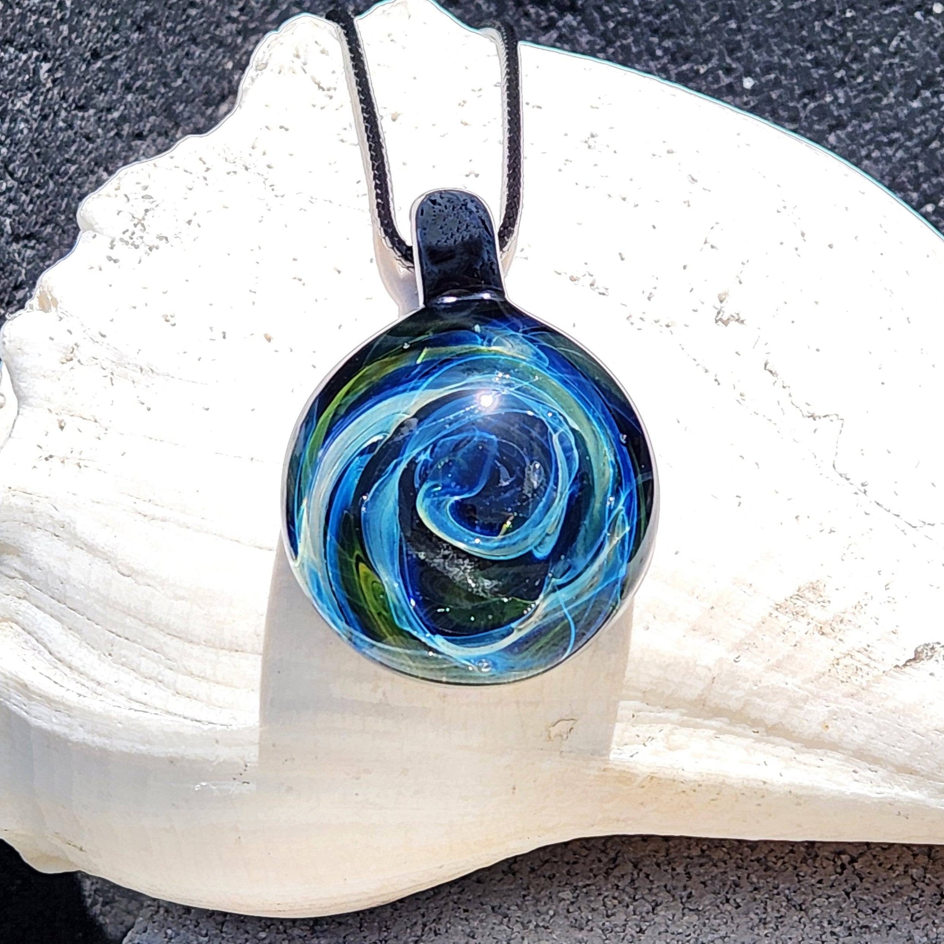 Heady Glass Pendant with swirling galaxy design in blue, green, and gold tones, handmade from borosilicate glass, displayed on a white seashell.