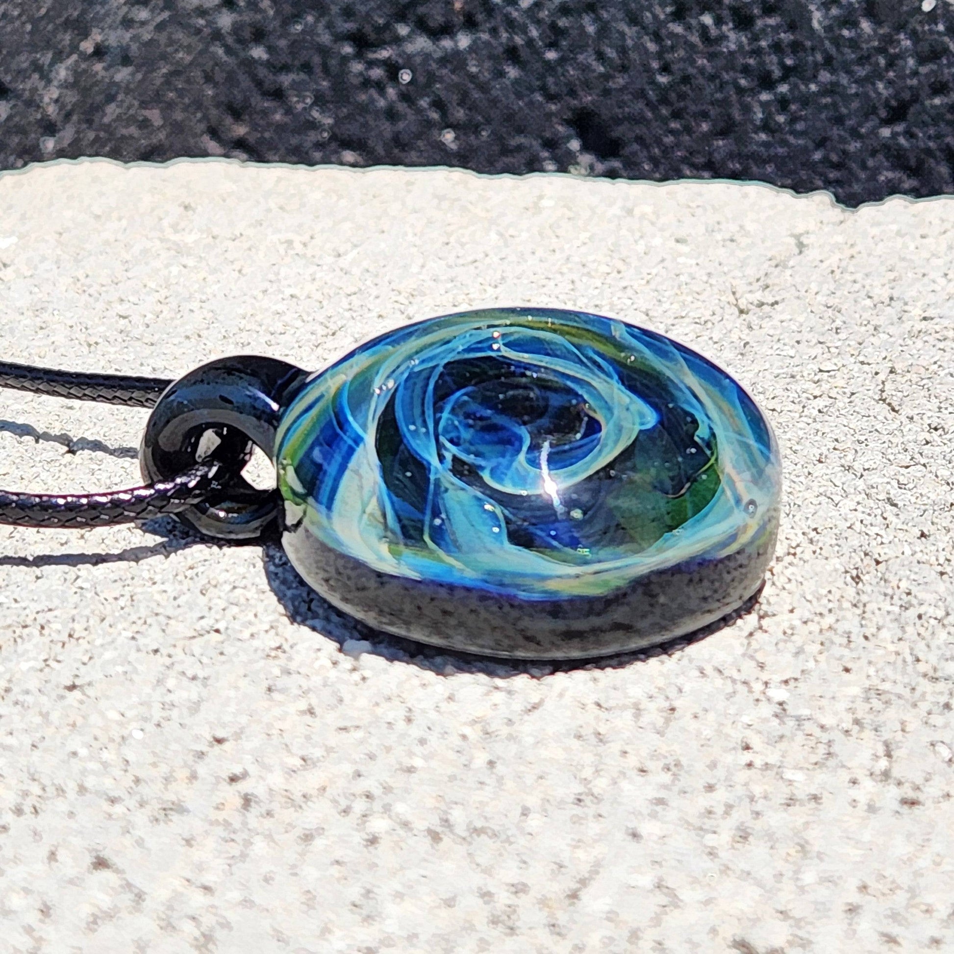 Artisan glass pendant with cosmic swirl design in blue and green hues, handcrafted from borosilicate glass and laid on a stone surface.