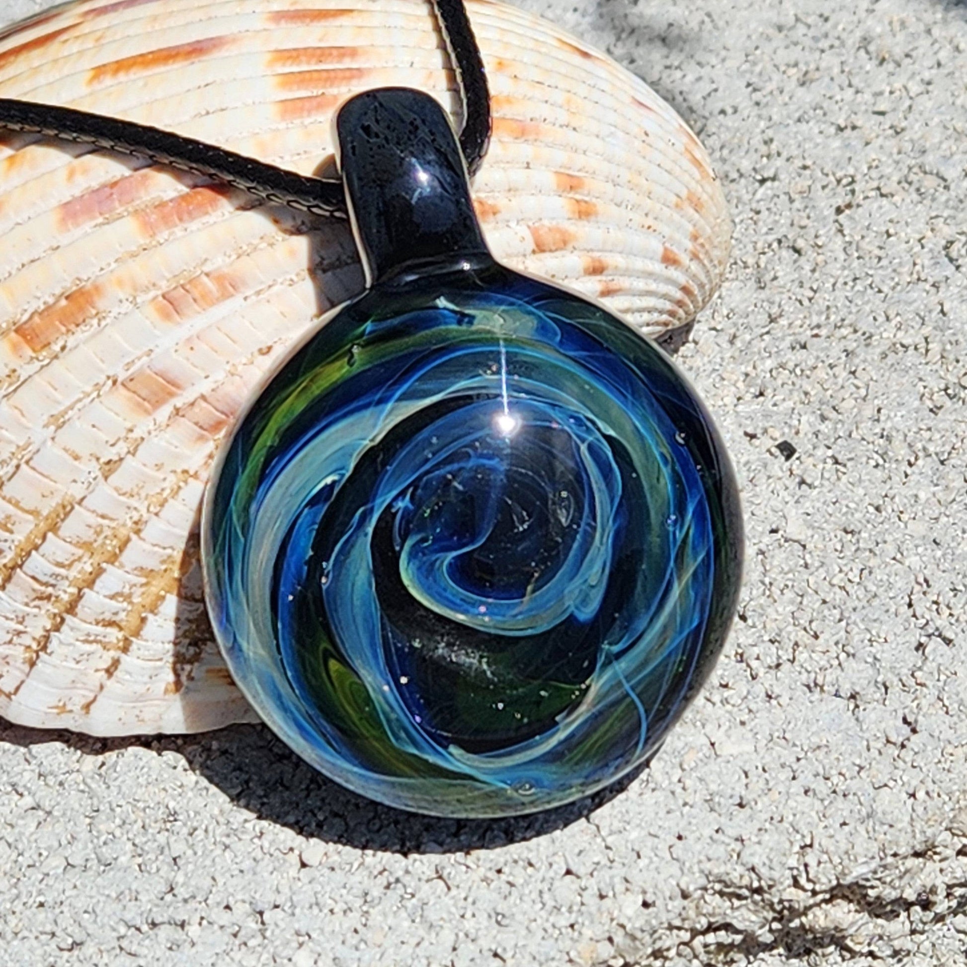 Galaxy-themed handmade glass pendant with vivid blue and green swirls, displayed against seashells and sand for a natural aesthetic.