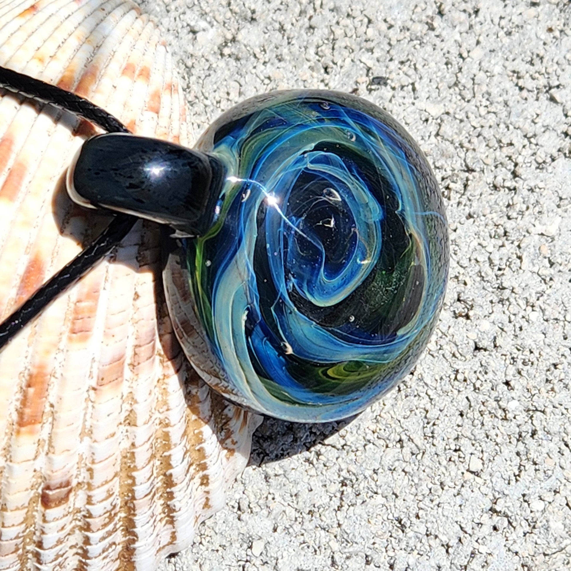 Close-up of handmade borosilicate glass pendant featuring a cosmic swirl pattern in blue and green, resting on a textured seashell.