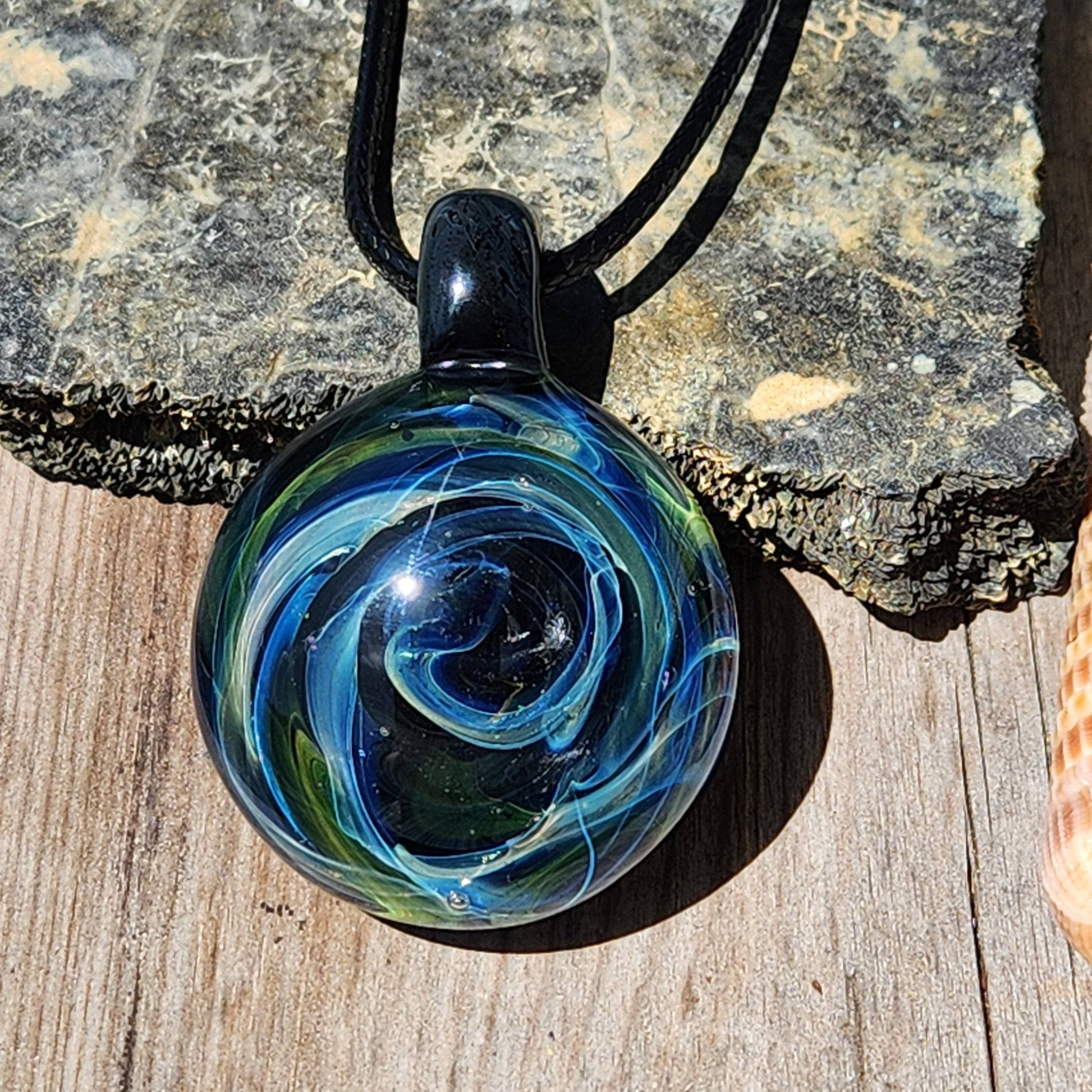 Handmade borosilicate glass pendant with swirling galaxy design in blue and green, photographed on rustic wood with natural lighting.