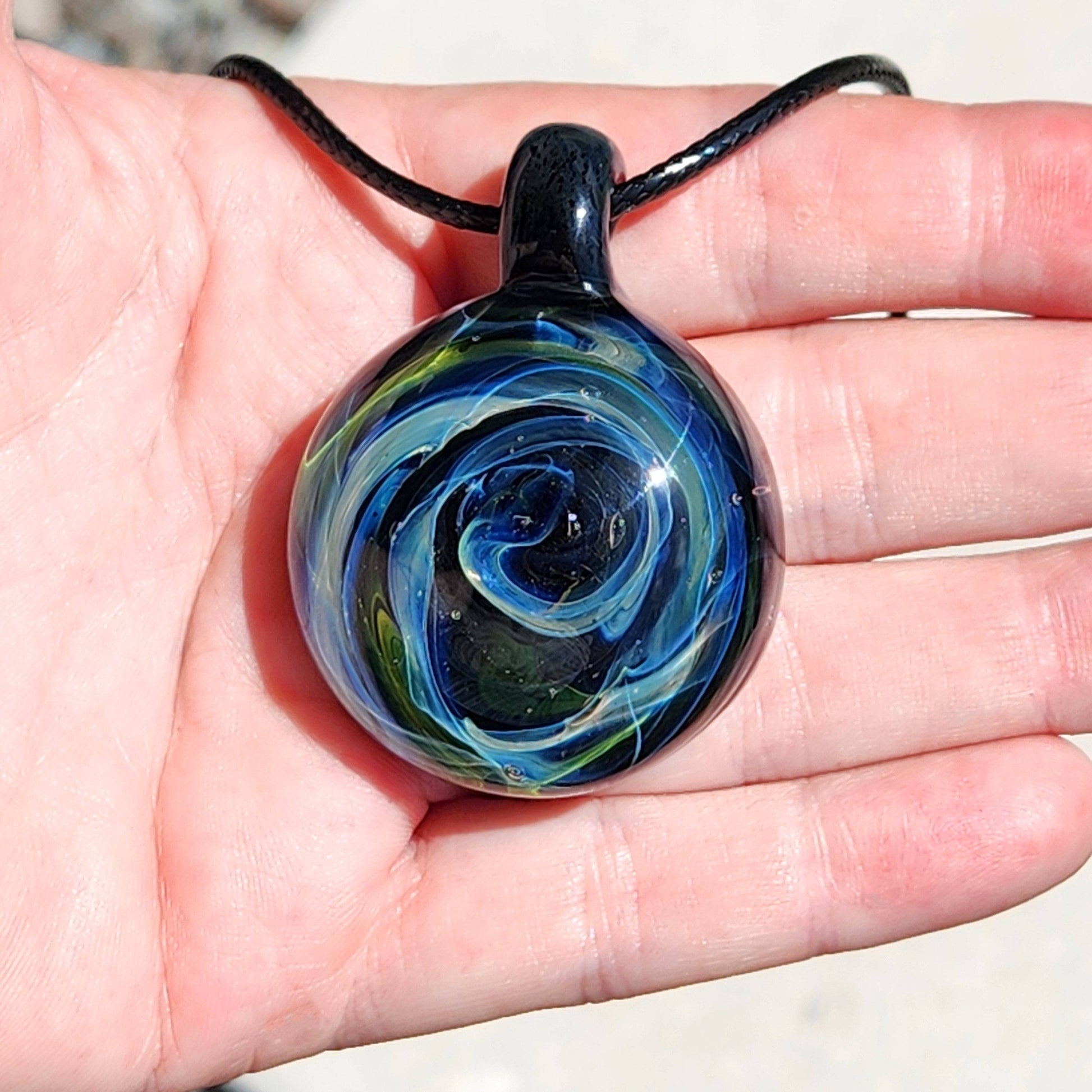 Handheld view of handmade borosilicate glass pendant with swirling blue and green galaxy pattern, showing its size and craftsmanship.