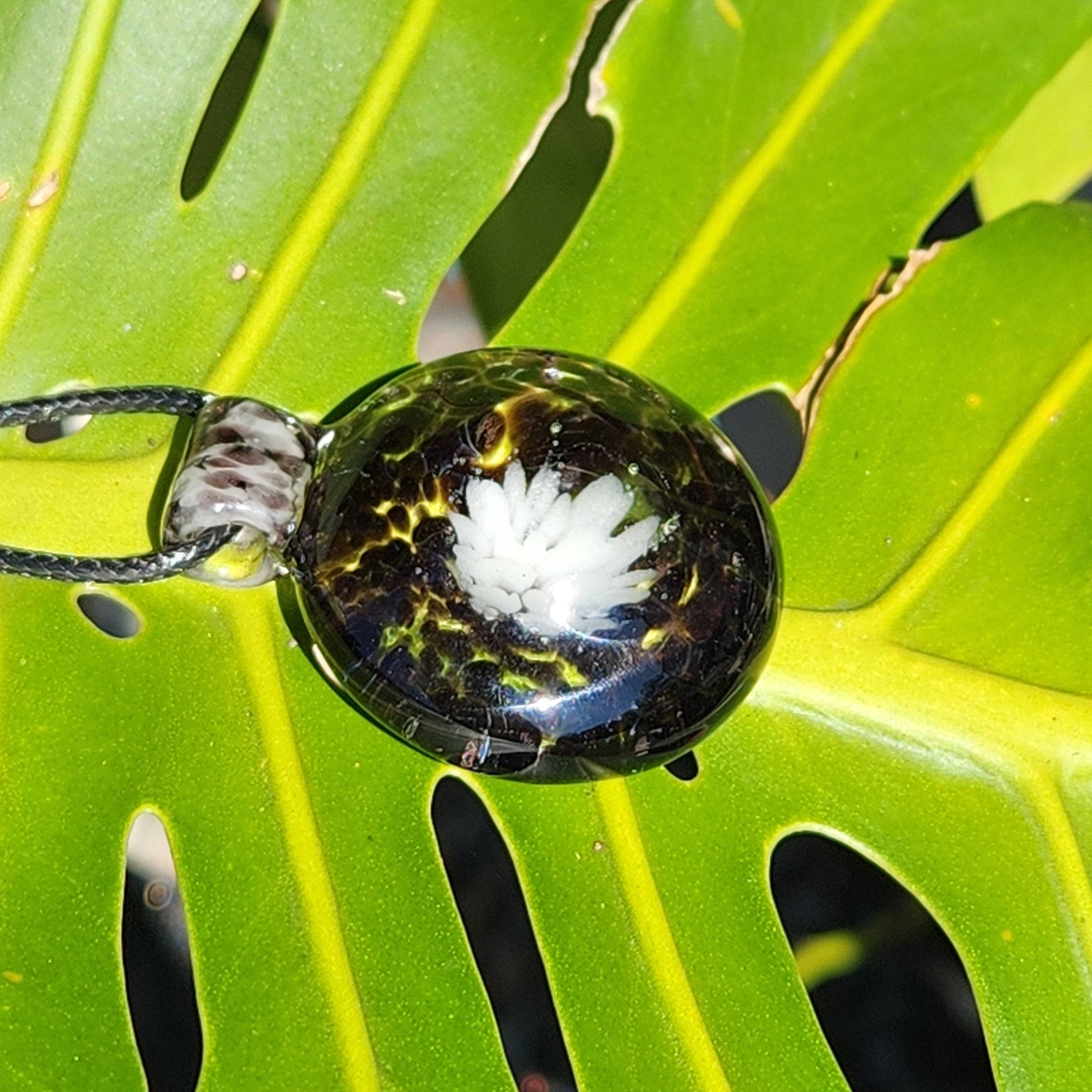 Heady Blown Glass Pendant resting on a green leaf, showcasing black and white floral design in a handmade borosilicate glass necklace.