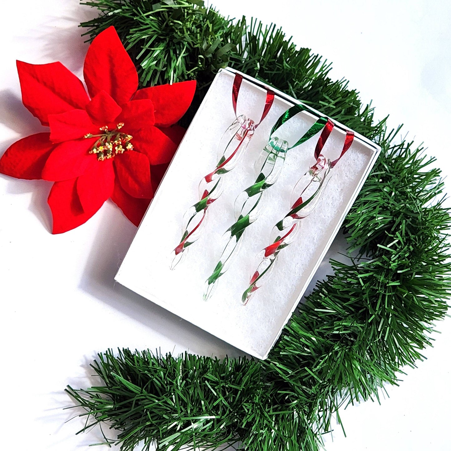 Gift box with three red and green handmade glass Christmas icicles, surrounded by garland and poinsettia for festive presentation.