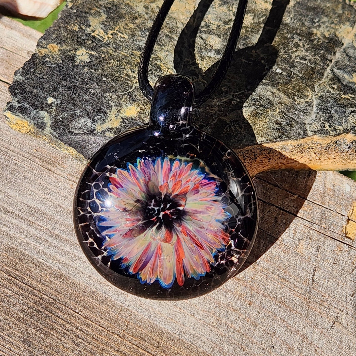 Handcrafted glass pendant with a vibrant flower design in red, purple, and cream, suspended on a black cord. One-of-a-kind jewelry piece.