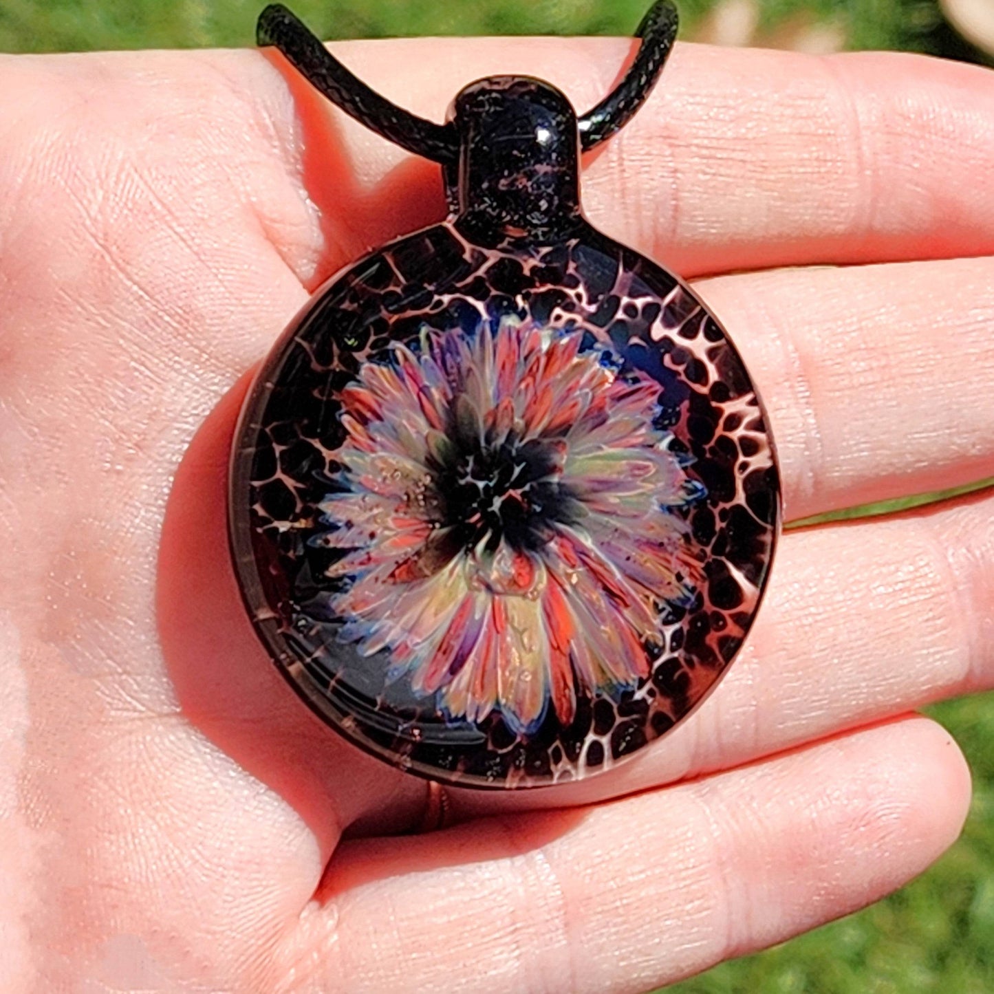 Handheld view of a handmade jewelry pendant with a vibrant flower design in purple, red, and cream, crafted from borosilicate glass.