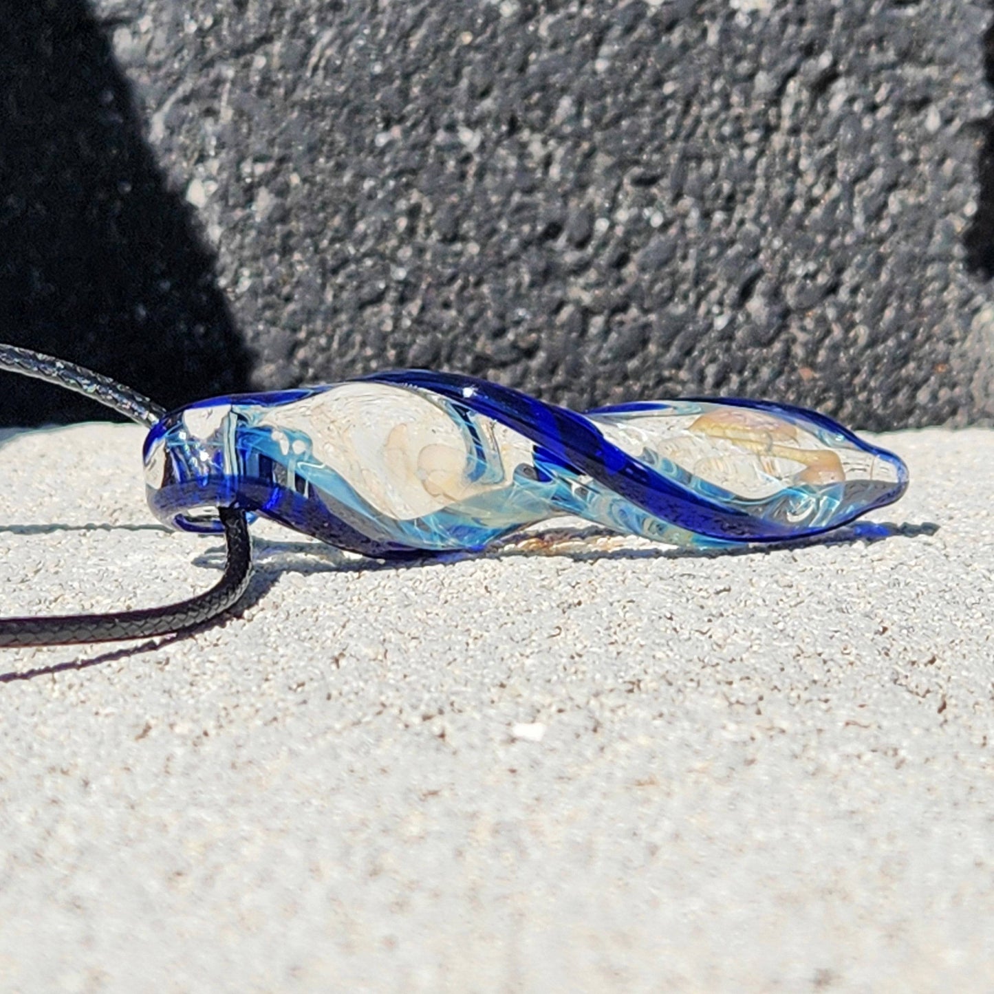 Close-up of handmade Blown Glass Spiral Pendant with silver-fumed core and blue spiral, lying flat on concrete surface.
