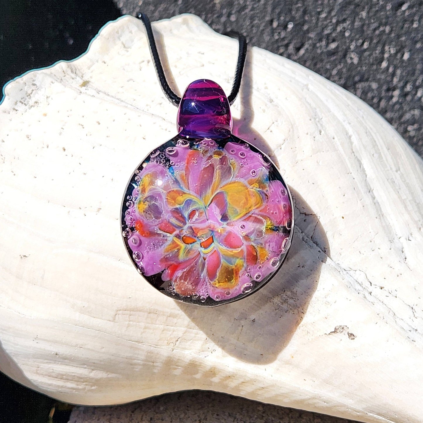 A handblown multicolored glass pendant with a floral pattern, attached to a black cord, displayed on a shell-shaped beaded necklace.