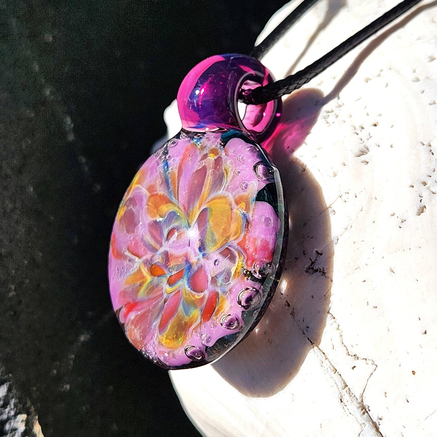 A handblown multicolored glass pendant with a floral pattern, attached to a black cord, displayed on a shell-shaped beaded necklace.