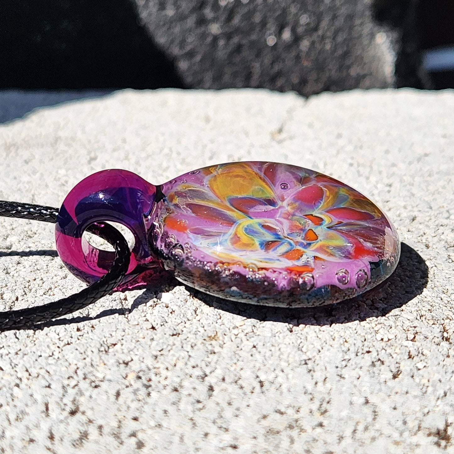 A side display of a handblown multicolored glass pendant with a floral pattern, attached to a black cord, displayed on a rock surface