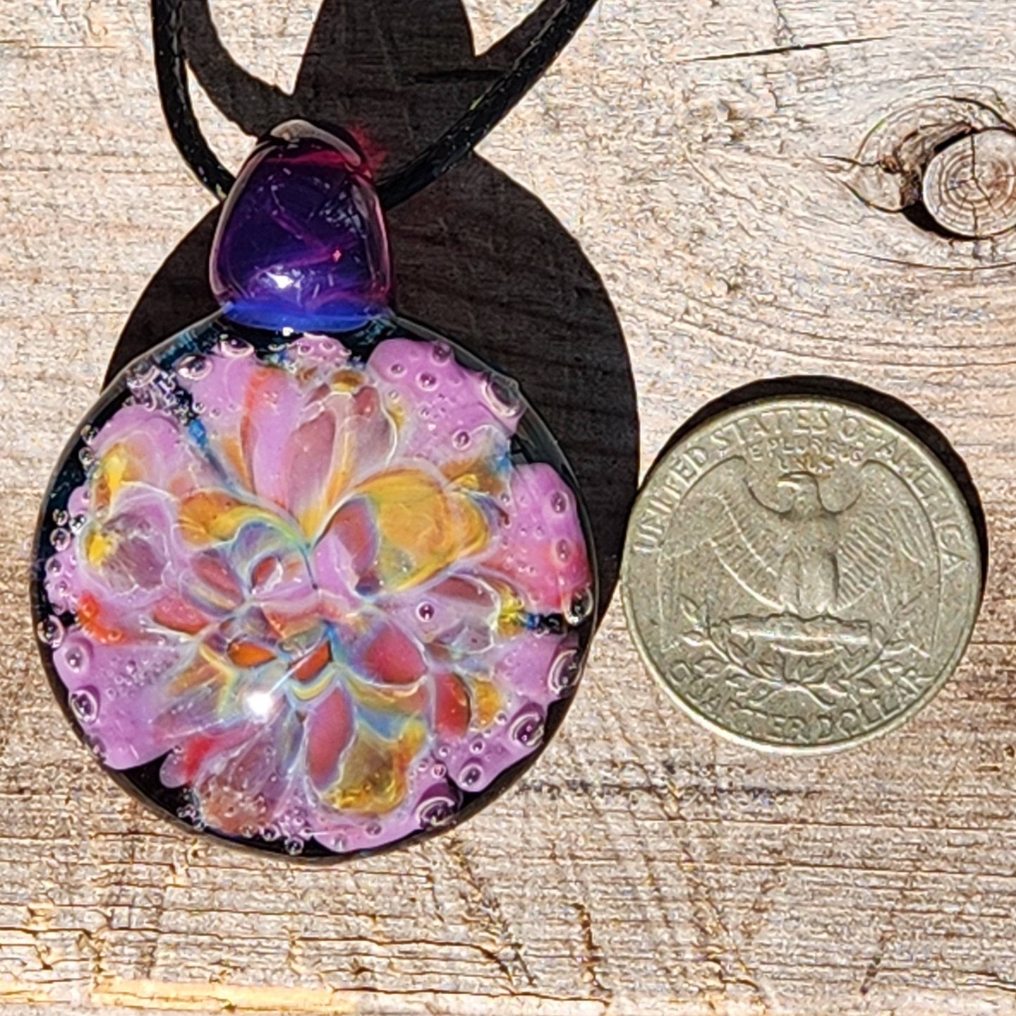 A handblown multicolored glass pendant with a floral pattern, attached to a black cord, displayed on a wood, next to a quarter for a size comparison.