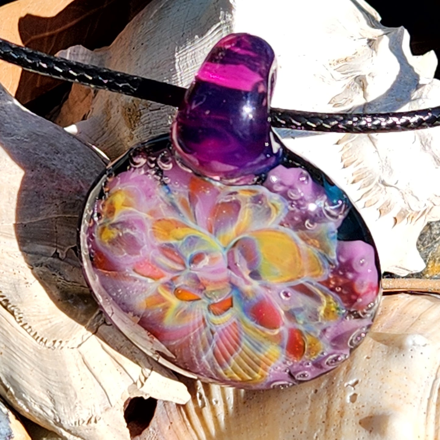 A handblown multicolored glass pendant with a floral pattern, attached to a black cord, displayed next to a shell-shaped beaded necklace.