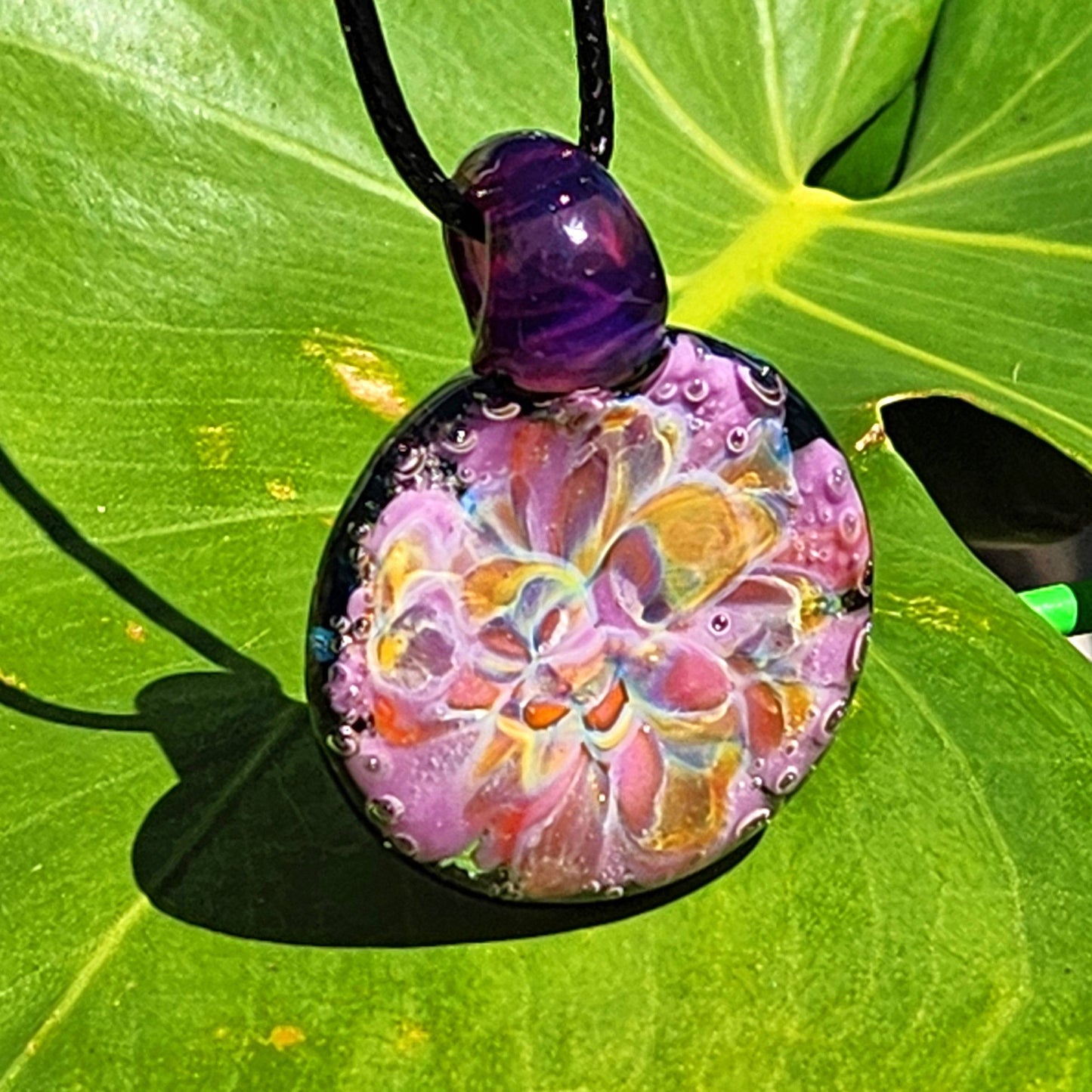 A handblown multicolored glass pendant with a floral pattern, attached to a black cord, displayed on a green leaf.