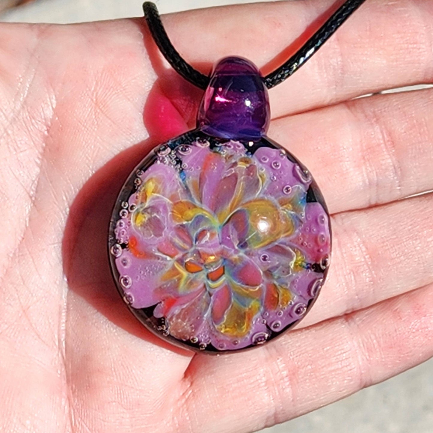 A handblown multicolored glass pendant with a floral pattern, attached to a black cord, displayed on a hand.