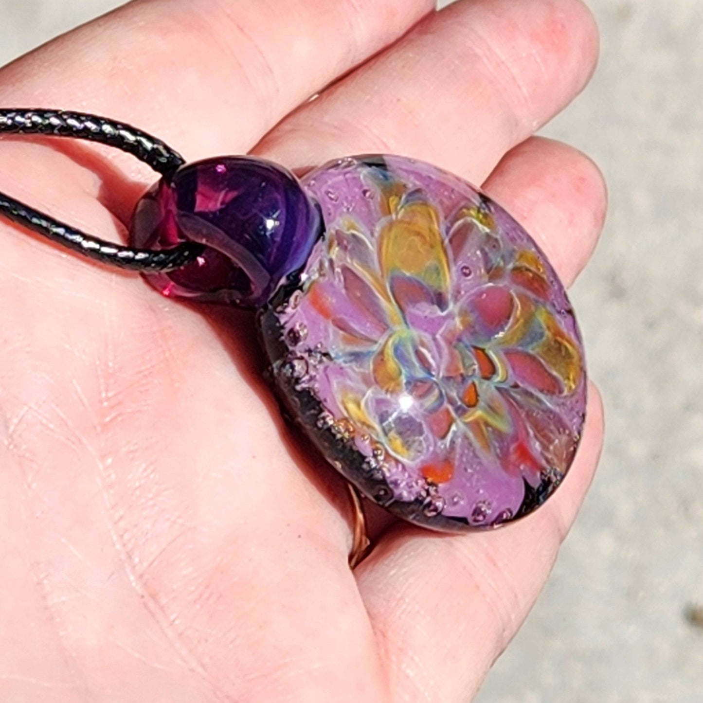 A hand displaying a handblown multicolored glass pendant with a floral pattern, attached to a black cord.