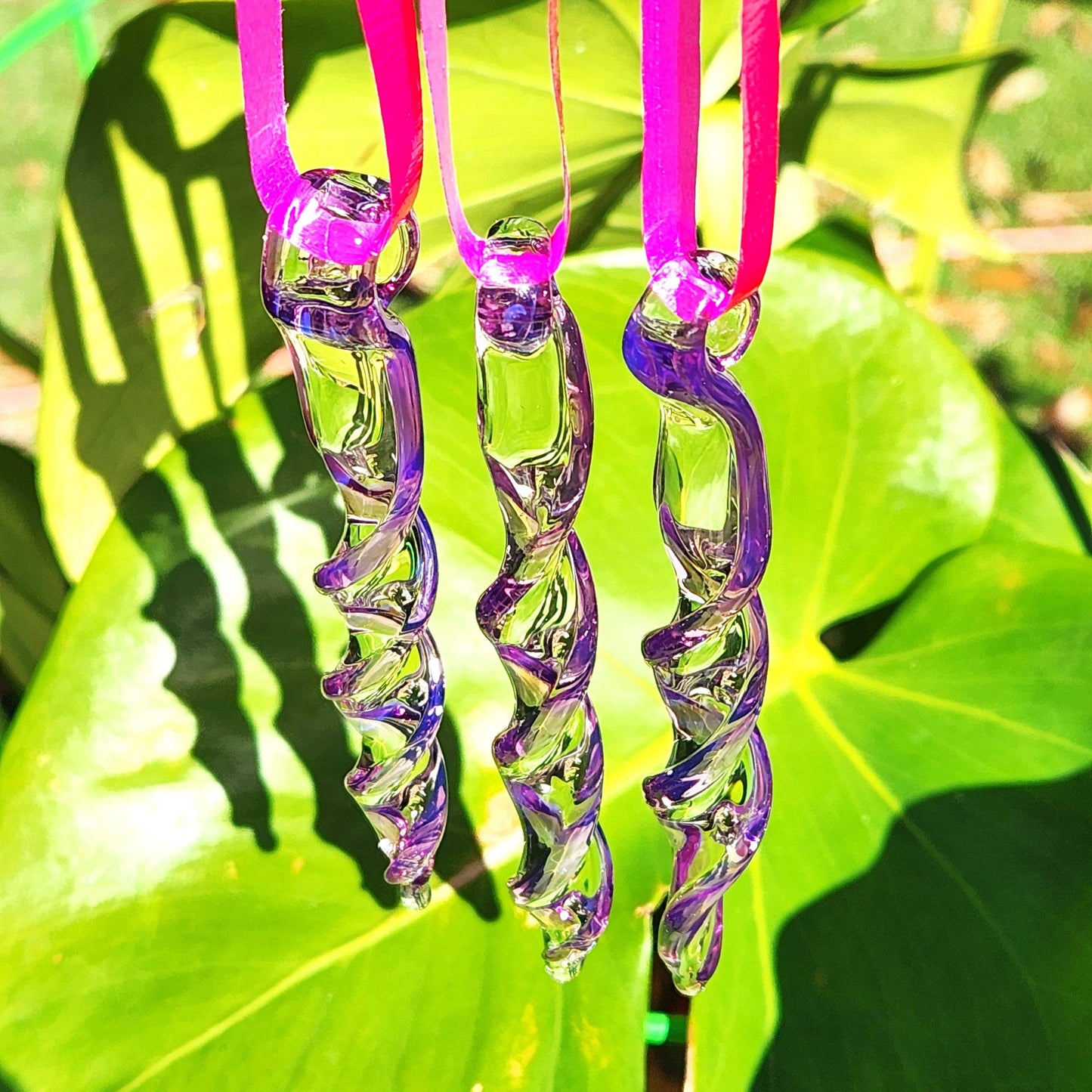 Three purple spiral glass suncatchers hanging in sunlight with green leaves in background, ideal for vibrant garden decor.