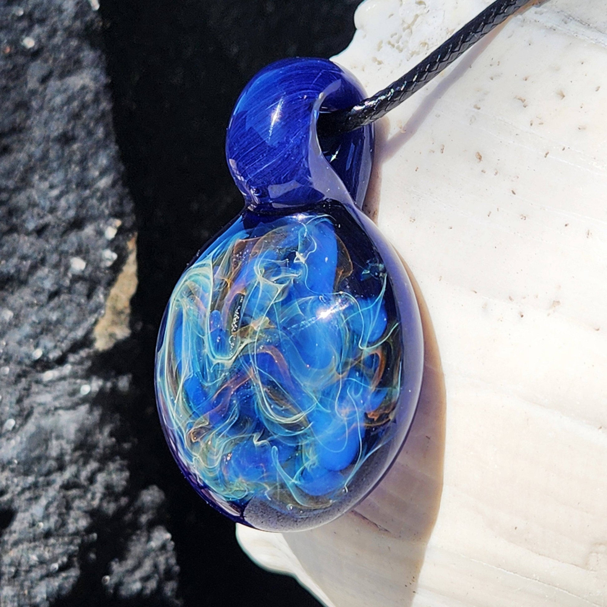 Angled view of handmade heady glass pendant with swirling blue and gold fumed accents, hanging on a black cord.