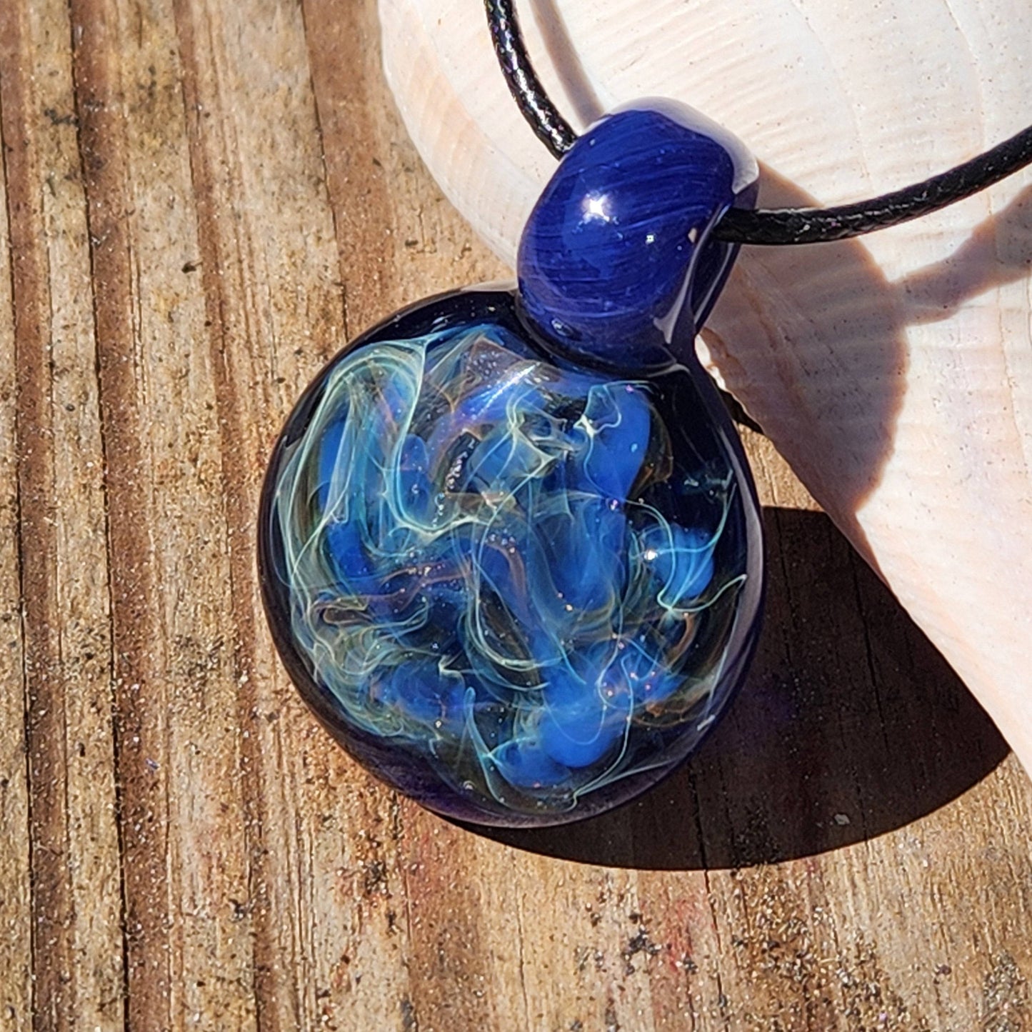 Handmade glass pendant with blue, silver, and gold fumed design resting on wood, perfect for gifting or personal wear.