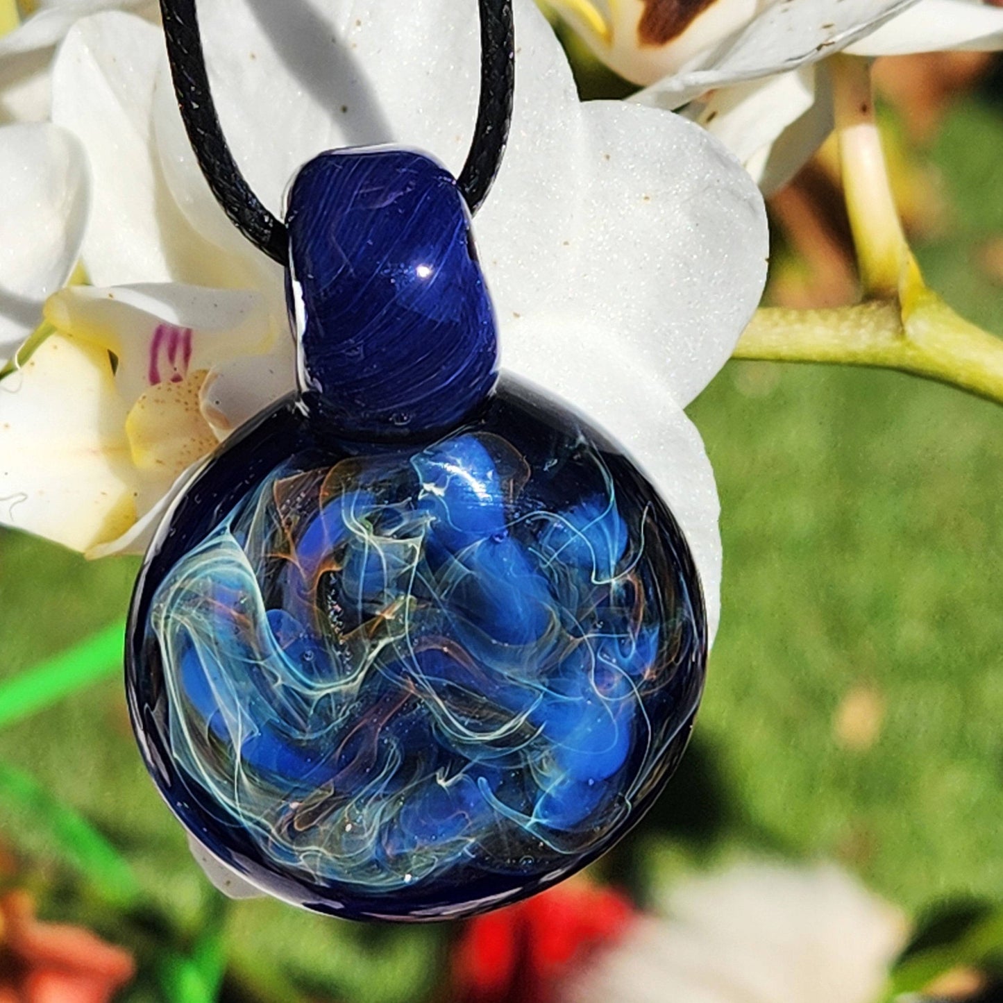 Trippy handmade glass pendant necklace with silver and gold fumed blue design, hanging in front of white orchid flowers.