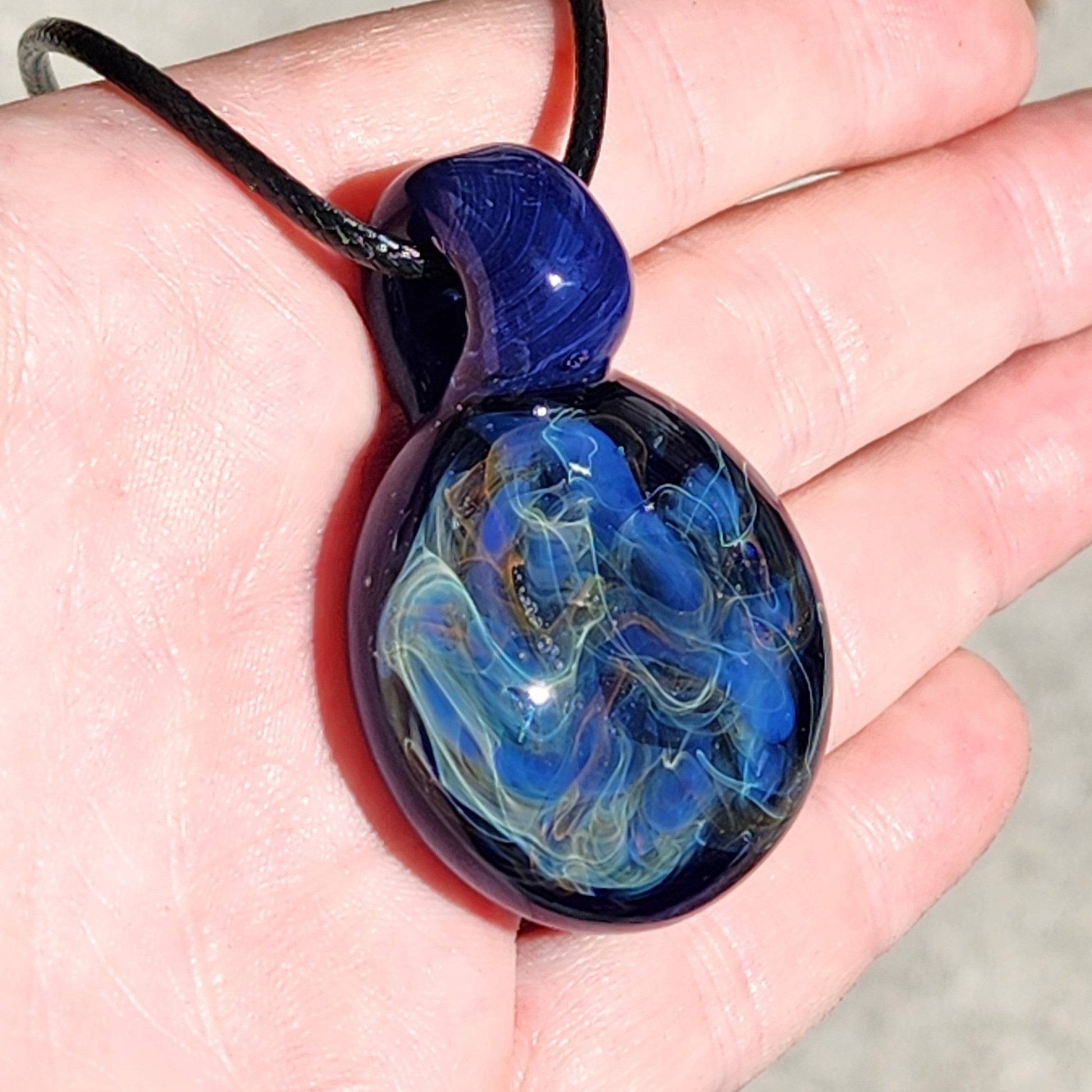 Hand holding a handmade heady glass pendant with vibrant blue and gold fumed swirls, highlighting its compact size and detail.