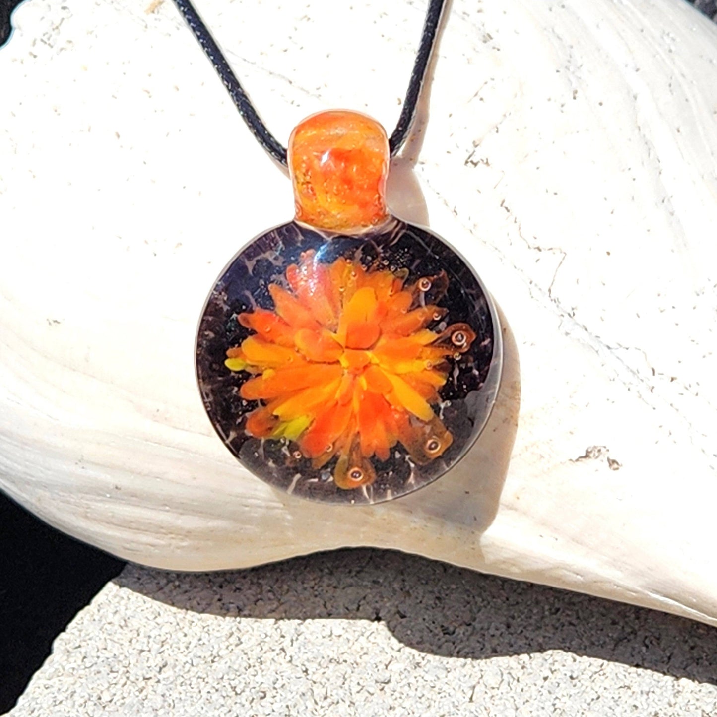 Borosilicate blown glass pendant necklace with vibrant red, yellow, and orange flower design, displayed on a white seashell background.
