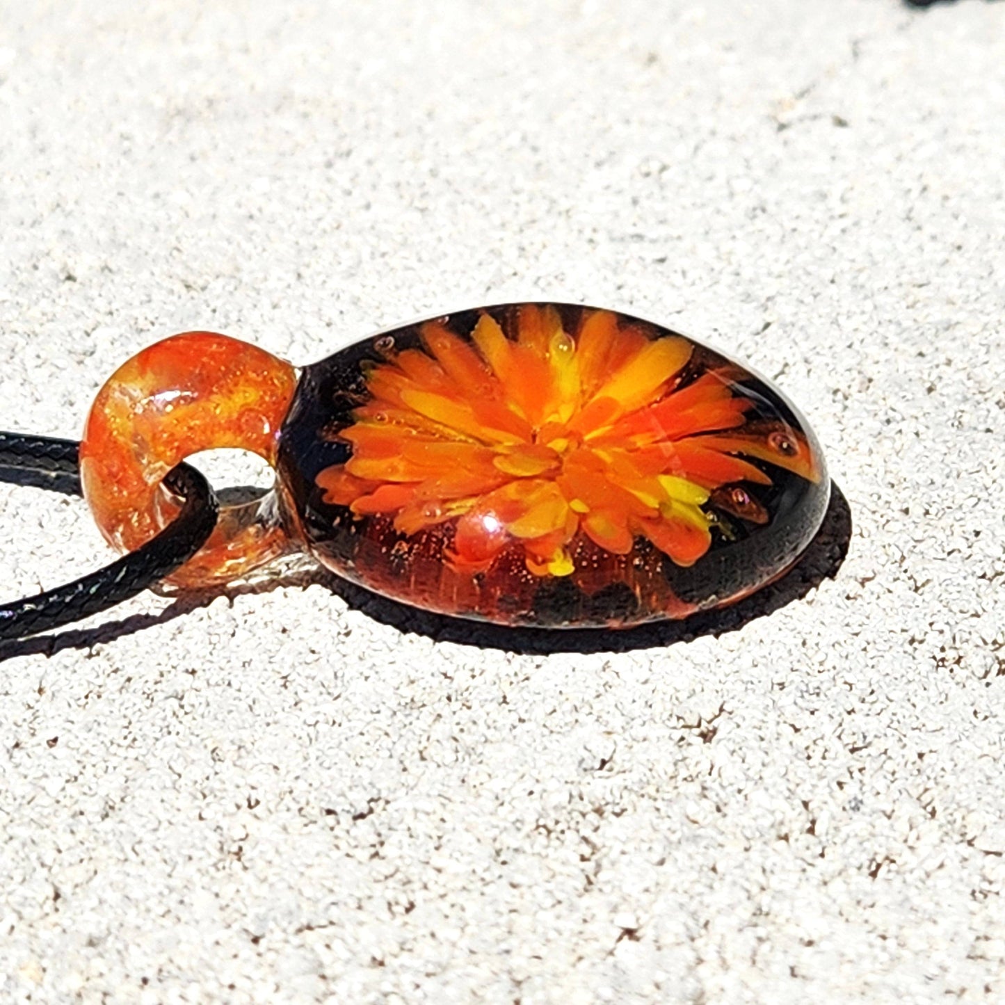 Flat view of borosilicate glass pendant necklace with vivid orange flower design, lying on sandy surface under sunlight.