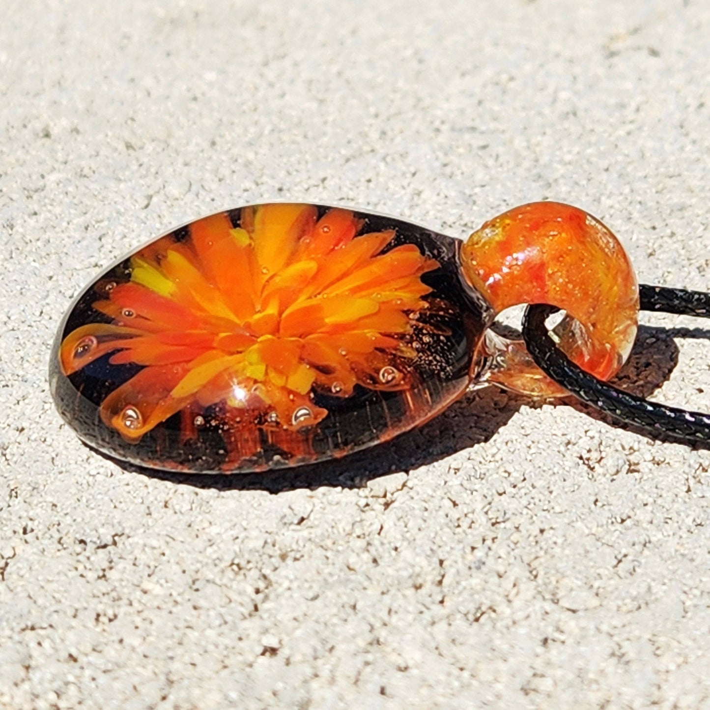 Side profile of handmade glass pendant necklace with orange and yellow flower design, lying on sand with black cord.
