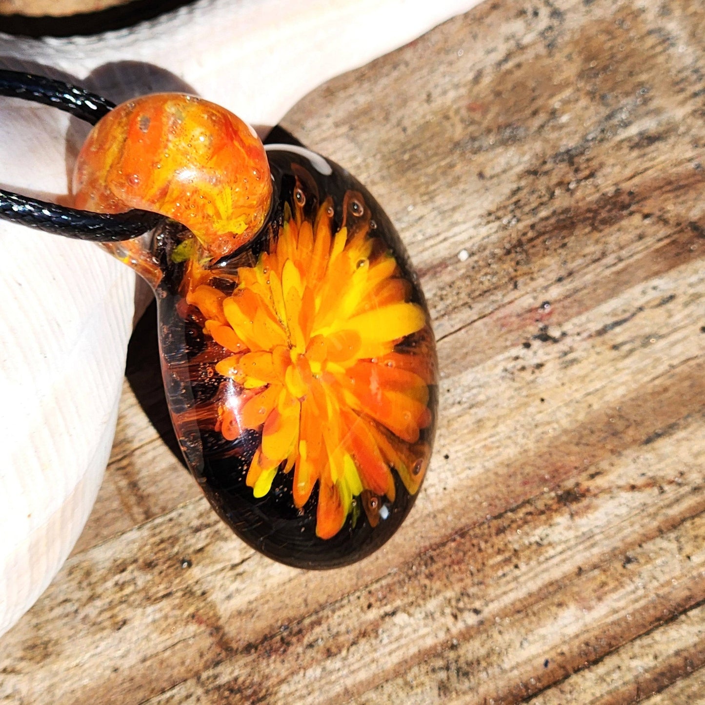 Angled view of handmade glass pendant necklace with swirling red, yellow, and orange flower, resting on wood and shell.