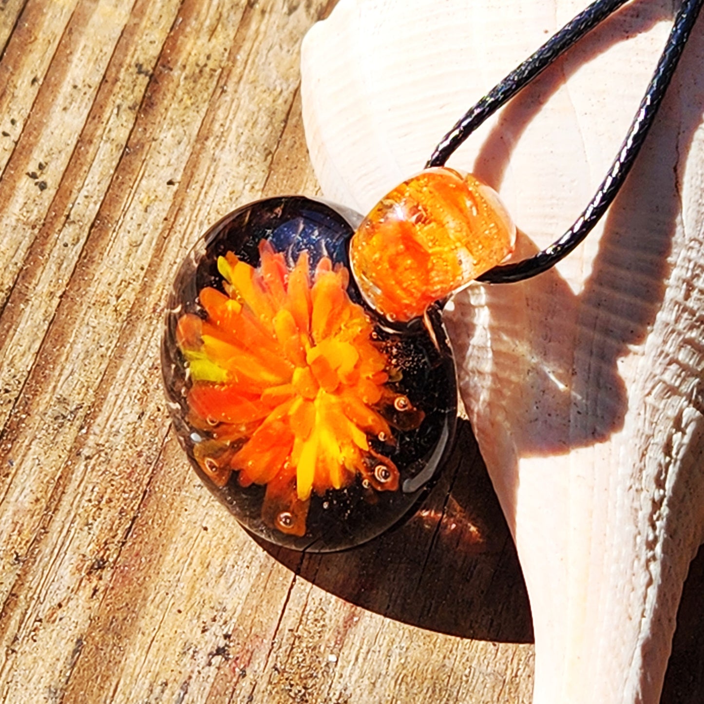 Artisan glass pendant necklace with swirling orange flower design, displayed on a shell and wooden surface in sunlight.