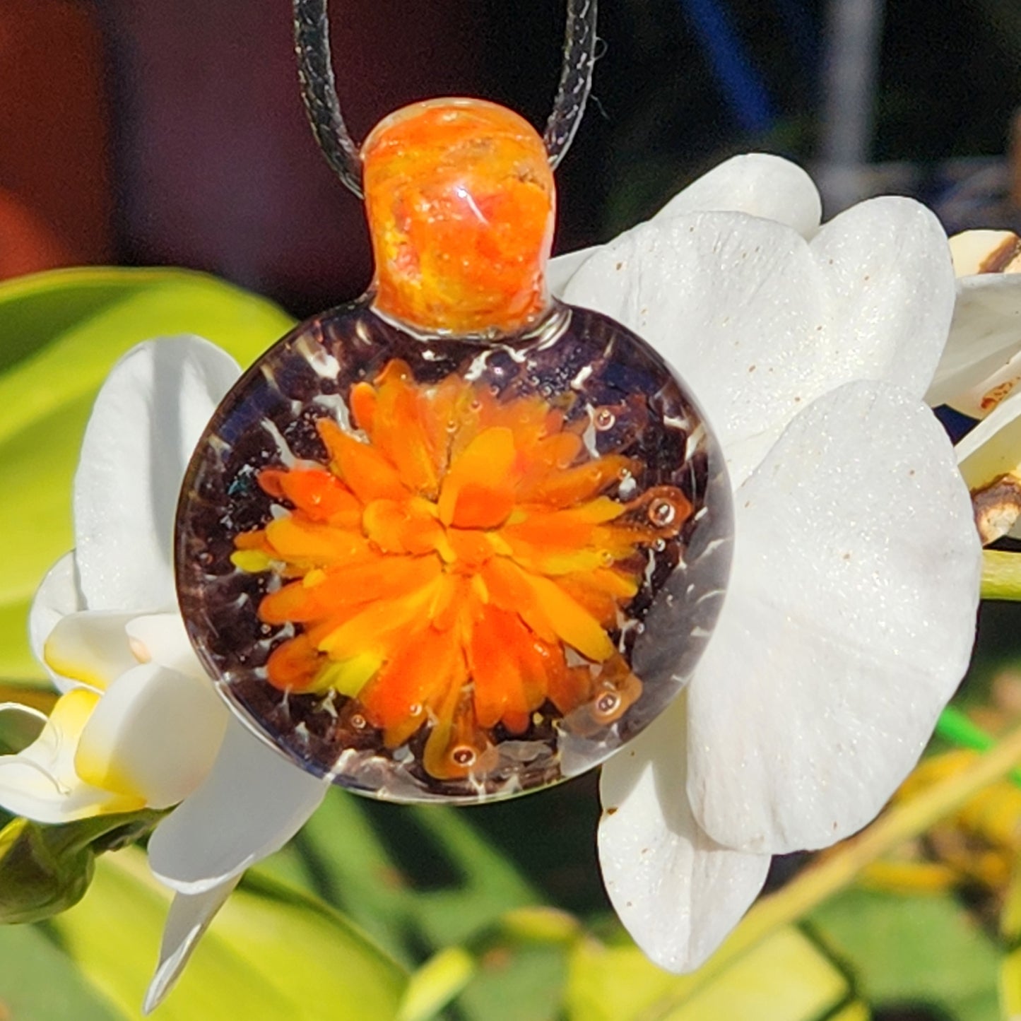 Handmade glass pendant necklace featuring fiery orange and yellow floral swirl, hanging in front of white tropical flowers.
