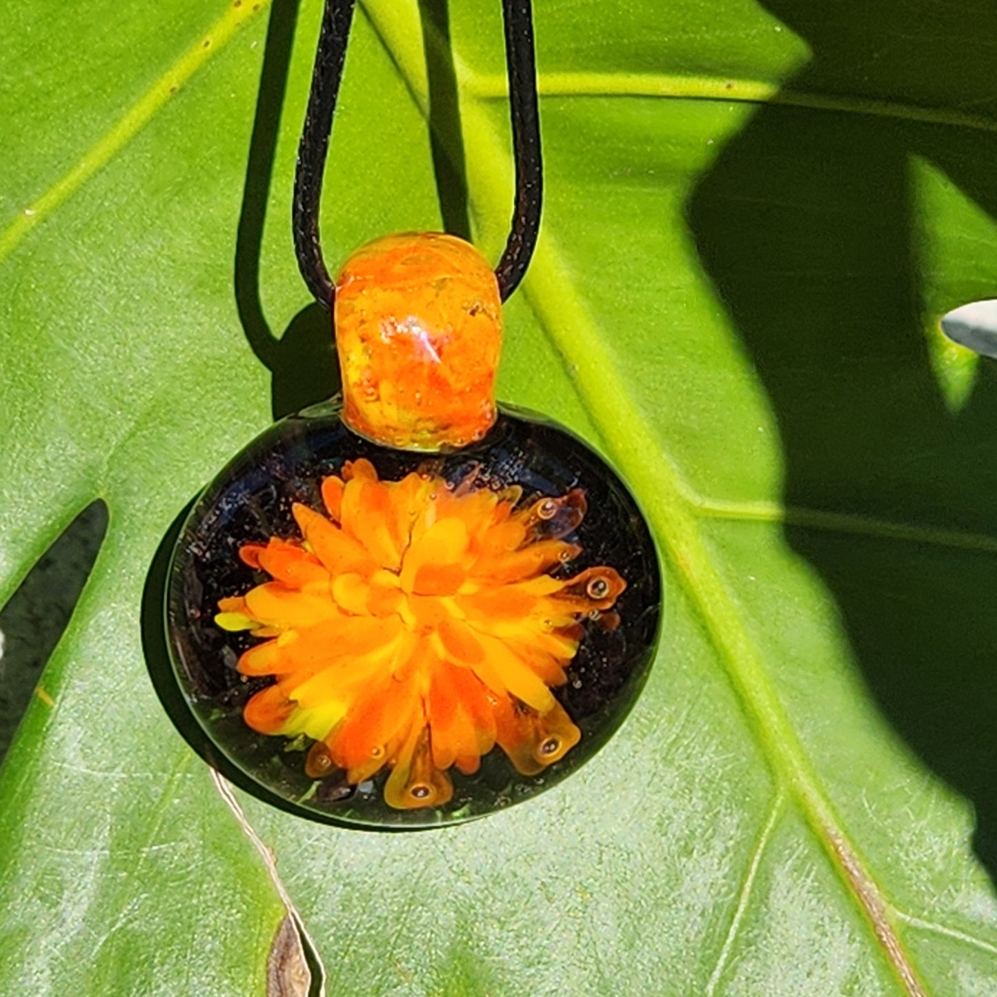 Borosilicate blown glass pendant necklace with fiery flower design, hanging against a green tropical leaf background.