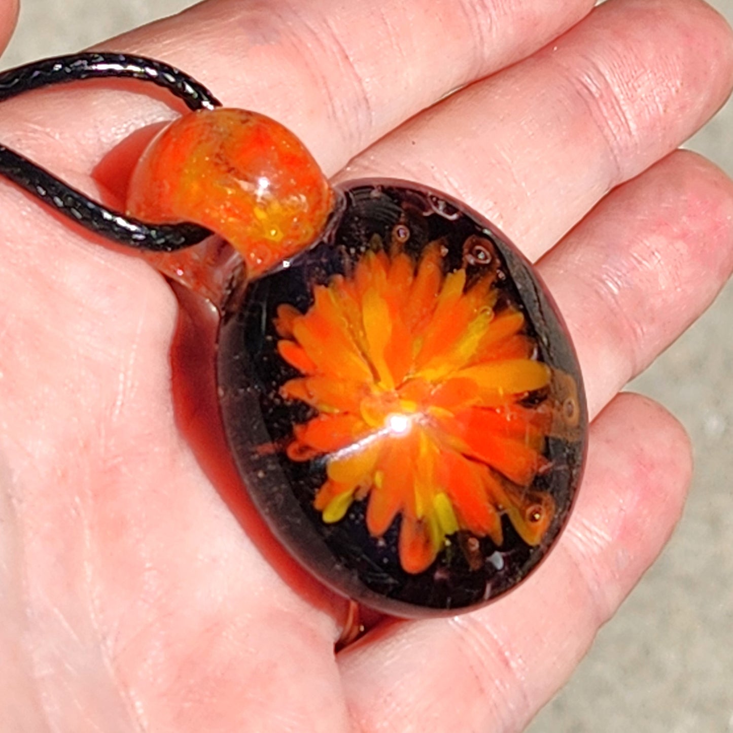 Hand holding borosilicate blown glass pendant necklace with vivid orange and yellow flower encased in clear glass.