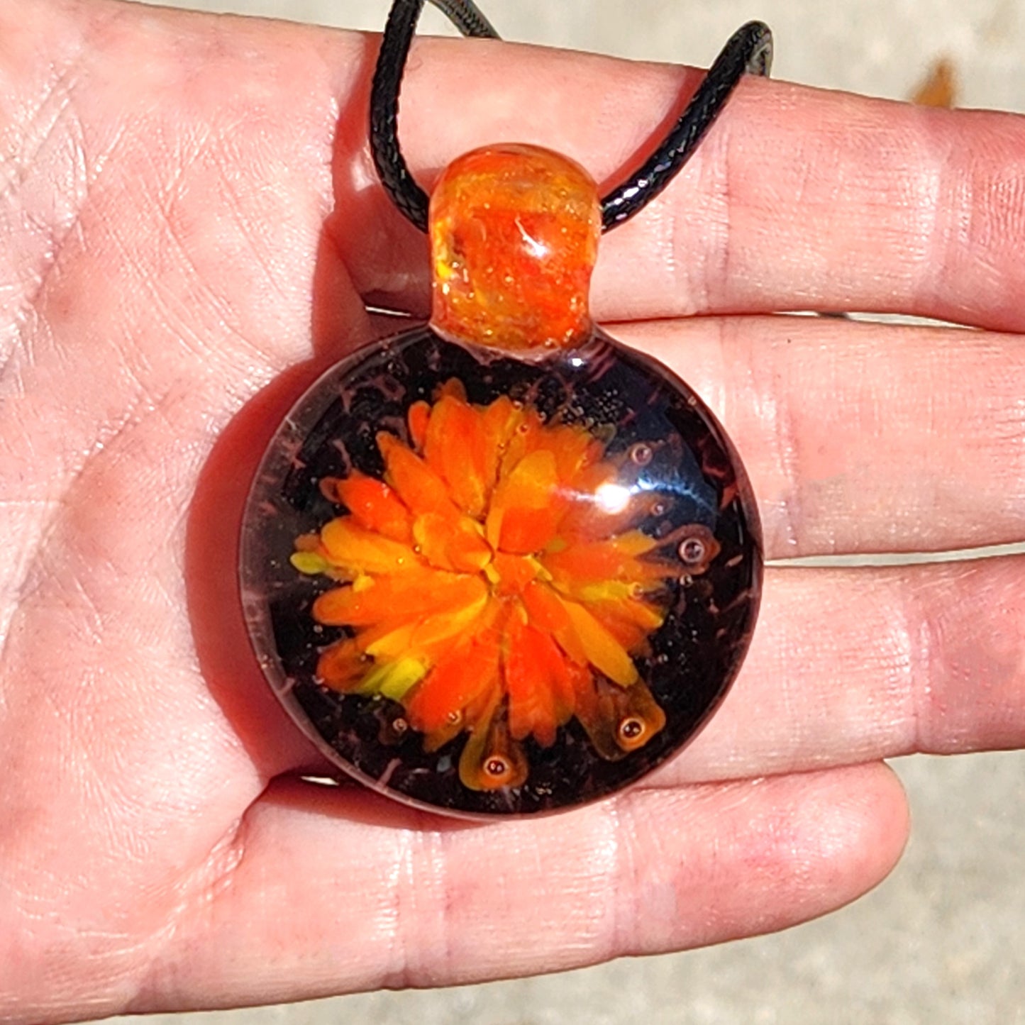 Close-up of handmade glass pendant necklace in hand, showcasing detailed red, orange, and yellow flower inside clear borosilicate glass.