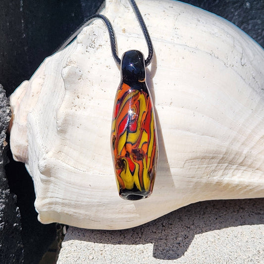 Handblown Tubular Glass Pendant with fiery red, orange, and yellow swirls on black, displayed on a seashell. Unique handmade jewelry gift.