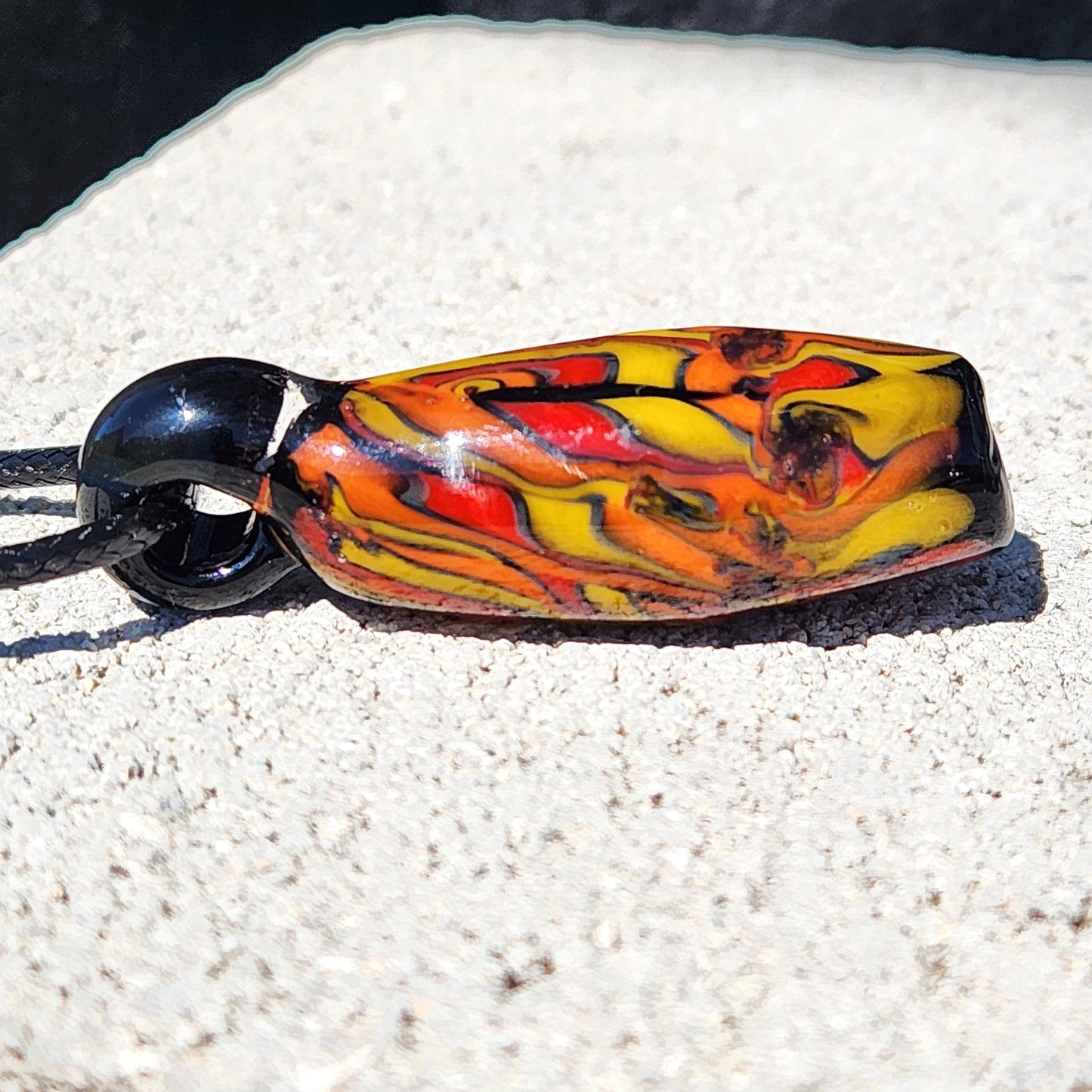 Side view of Handblown Tubular Glass Pendant with swirling flame colors on black, lying on stone surface. Unique handmade jewelry charm.
