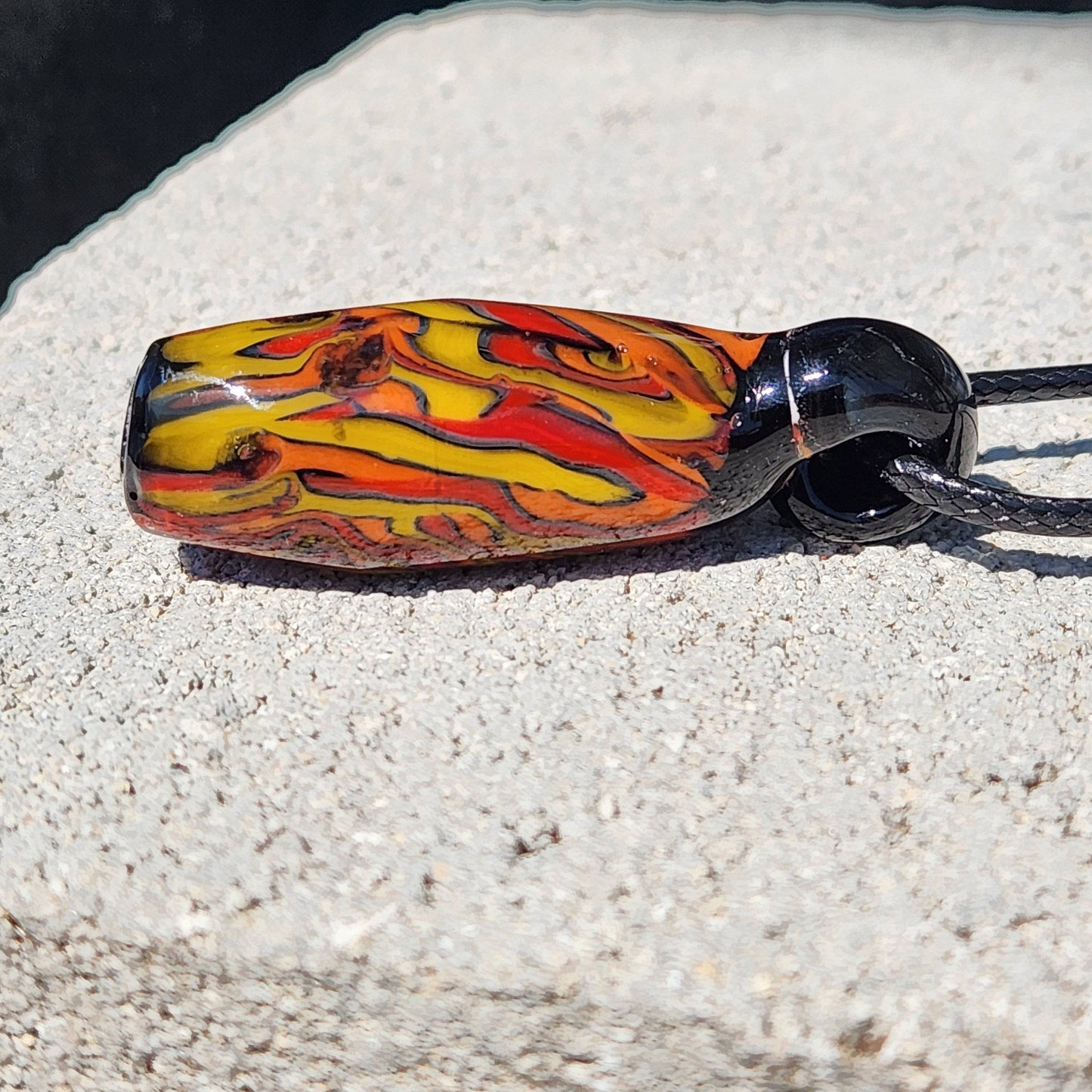 Handblown Tubular Glass Pendant with vibrant flame-like colors on black, lying horizontally on stone. Unique handmade jewelry piece.