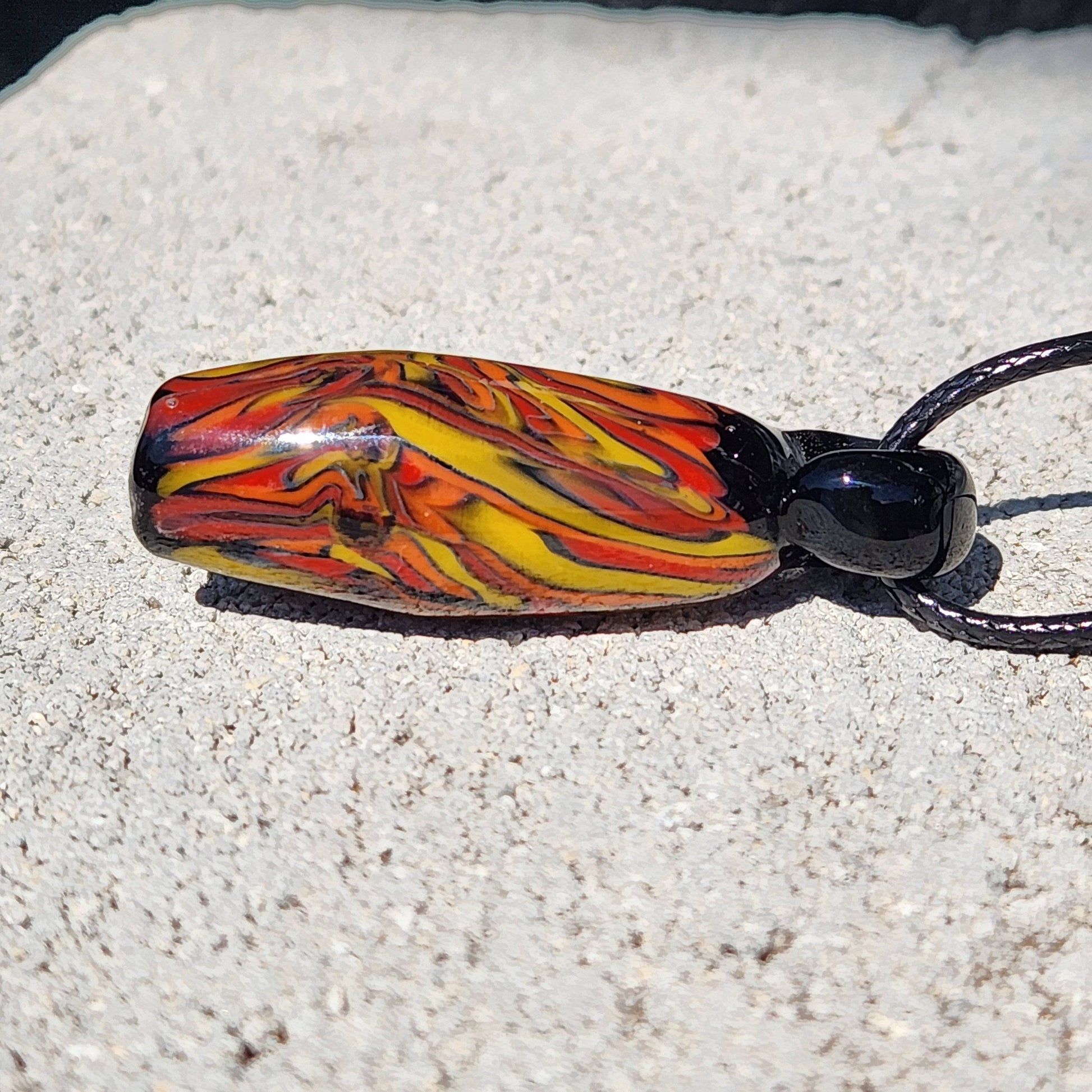 Fiery red, orange, and yellow Handblown Tubular Glass Pendant with black loop, displayed on stone. Handmade jewelry gift for unique style.