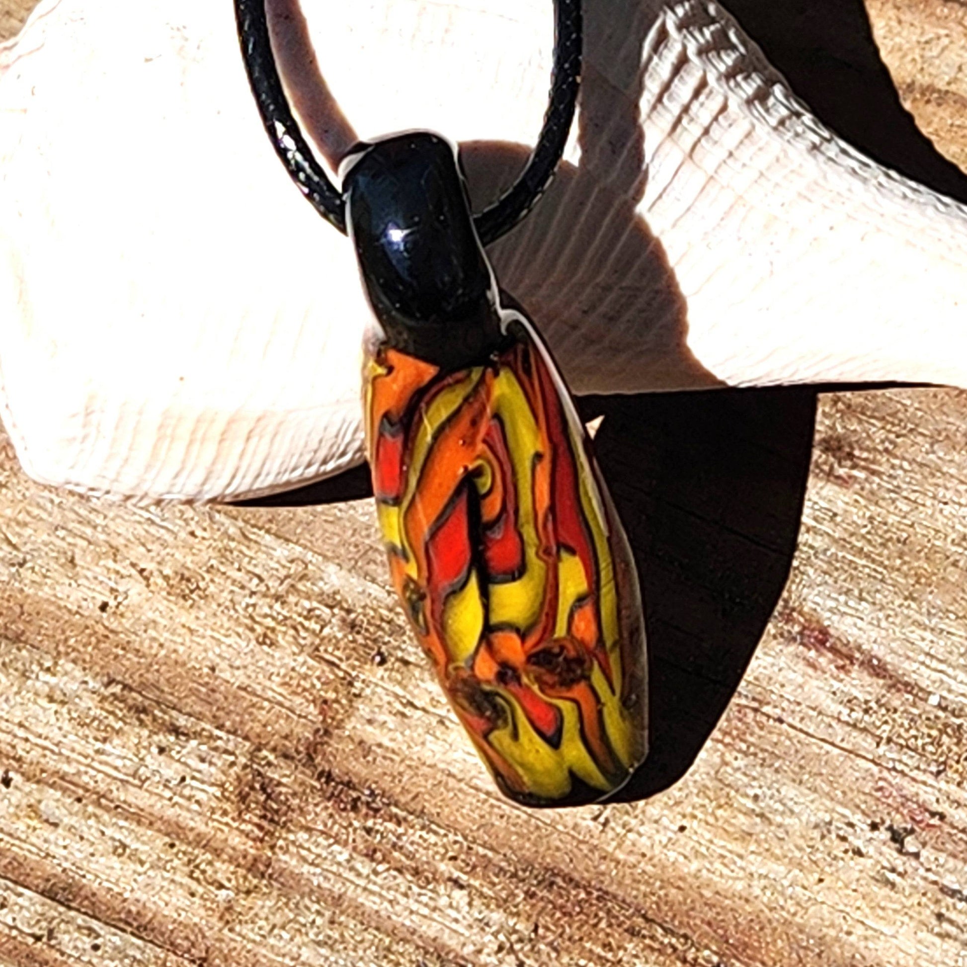 Handmade Glass Pendant with molten lava-inspired colors resting on wood and shell. Handblown tubular design in red, orange, and yellow tones.