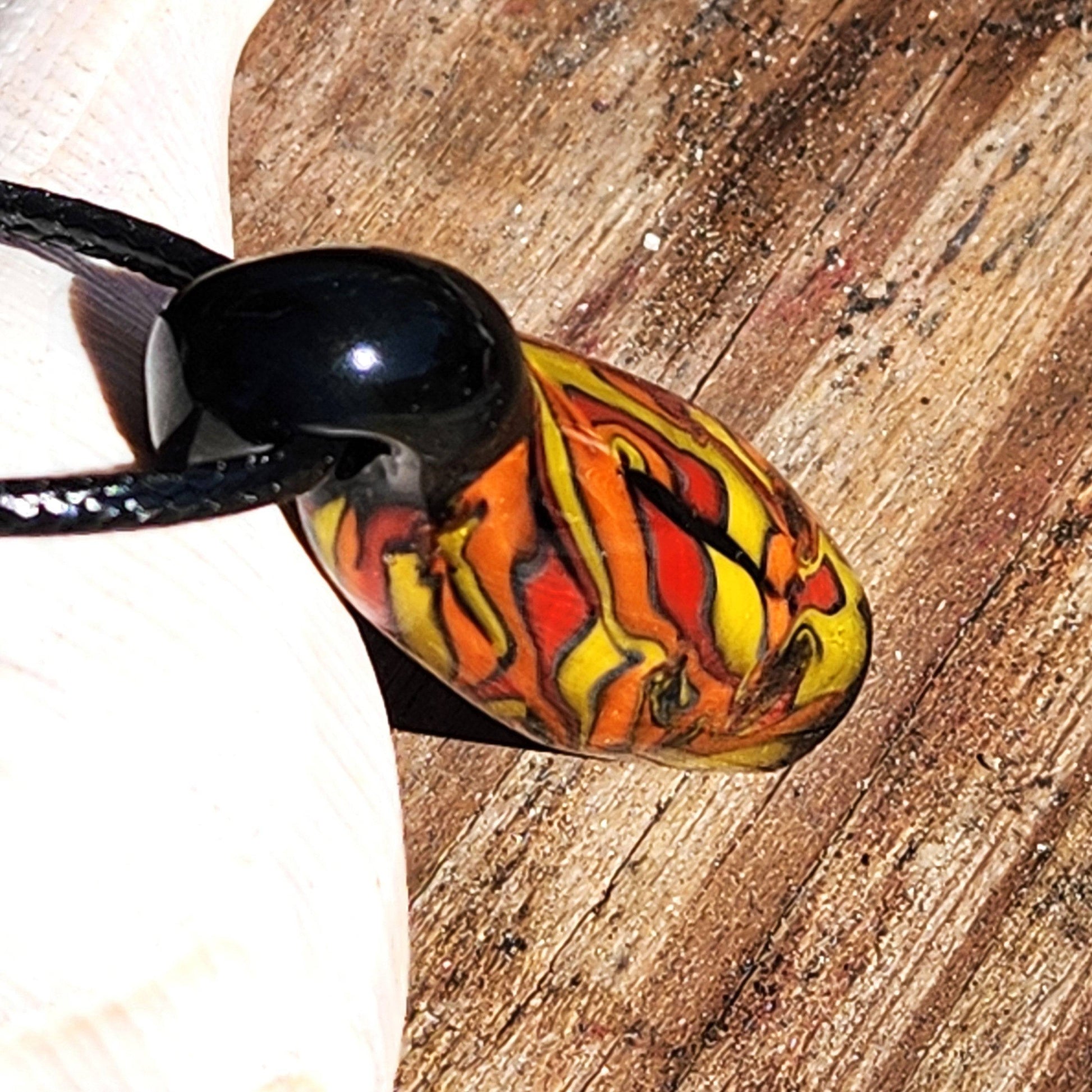 Close-up of Handblown Tubular Glass Pendant with bold orange, red, and yellow streaks, resting on wood. Handmade jewelry with fiery design.