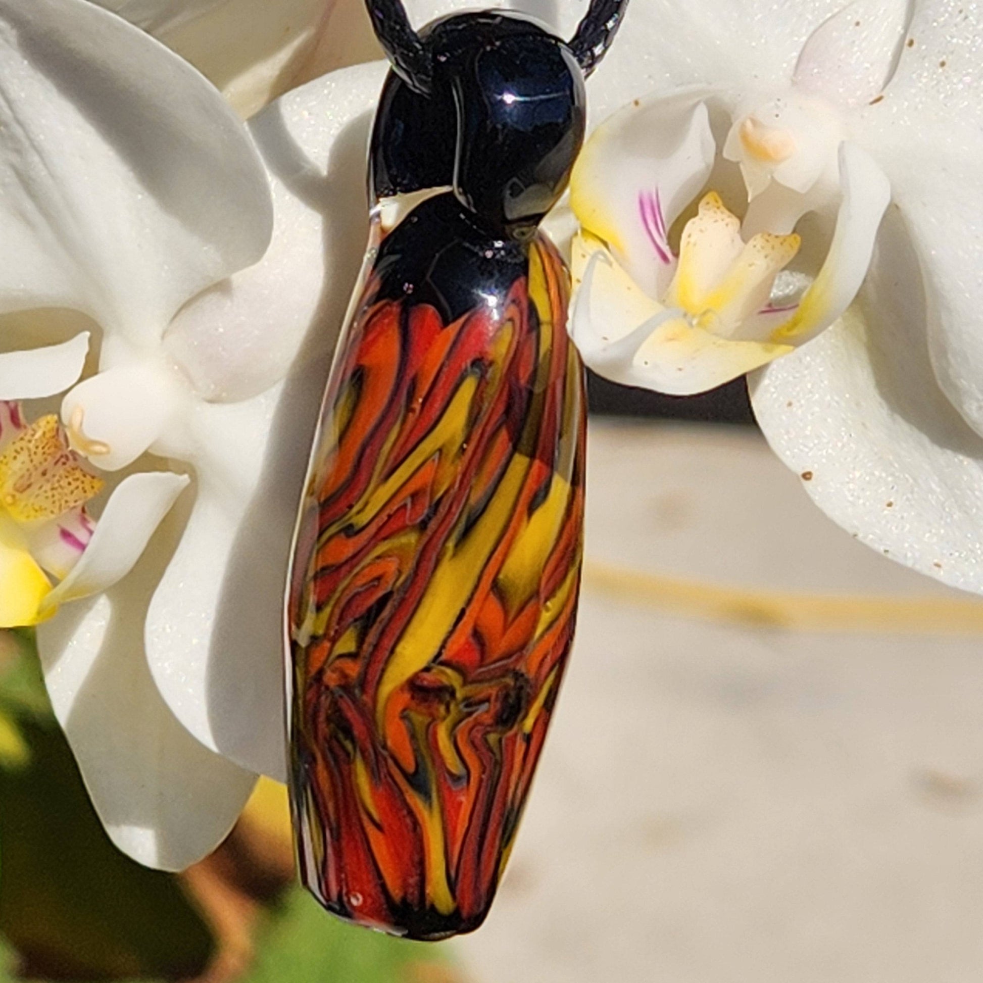 Close-up of Handblown Tubular Glass Pendant with vivid flame-like patterns, hanging in front of white orchid flowers. Handmade jewelry charm.