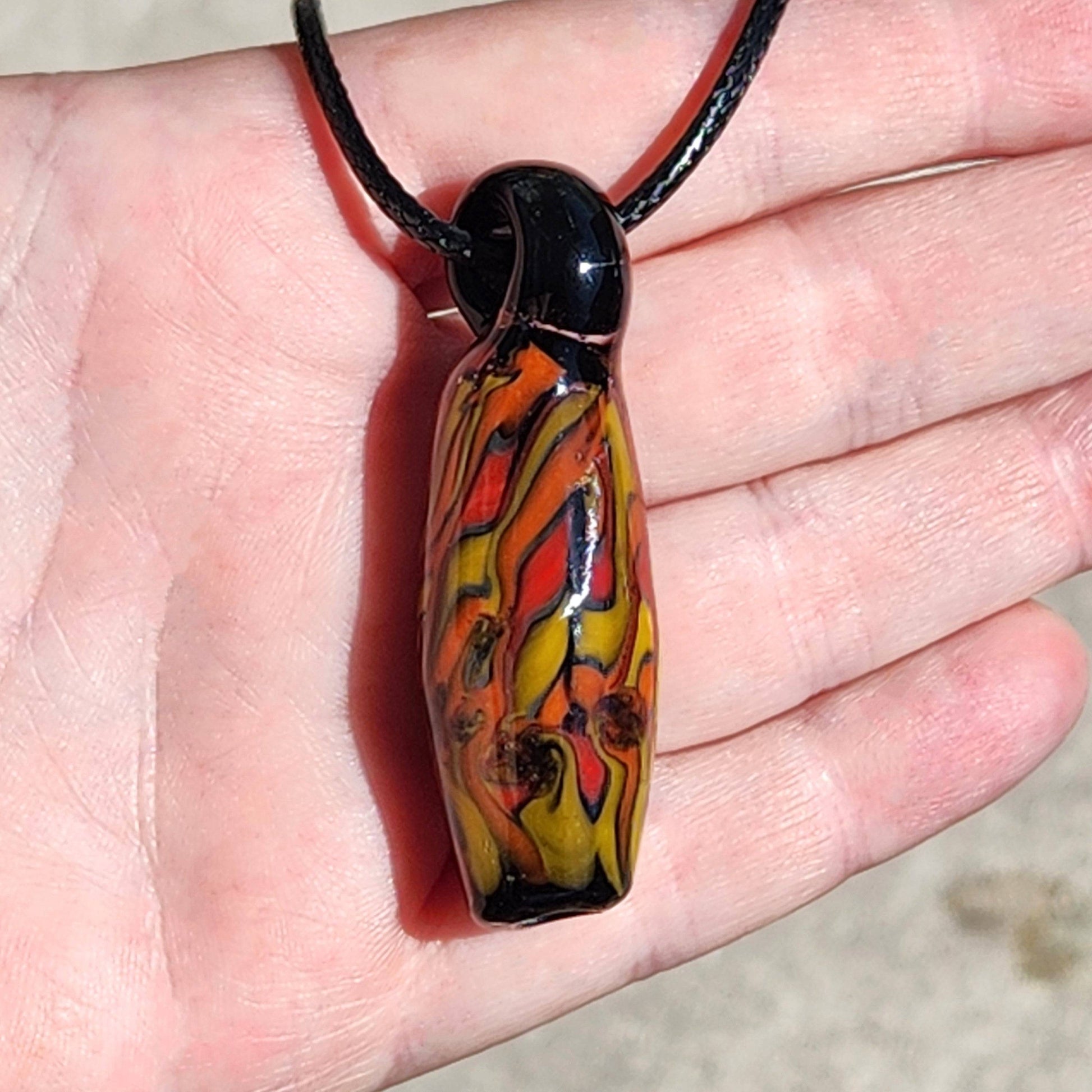 Handmade Tubular Glass Pendant with red, orange, and yellow flame design held in palm. Unique handblown jewelry gift with black loop.
