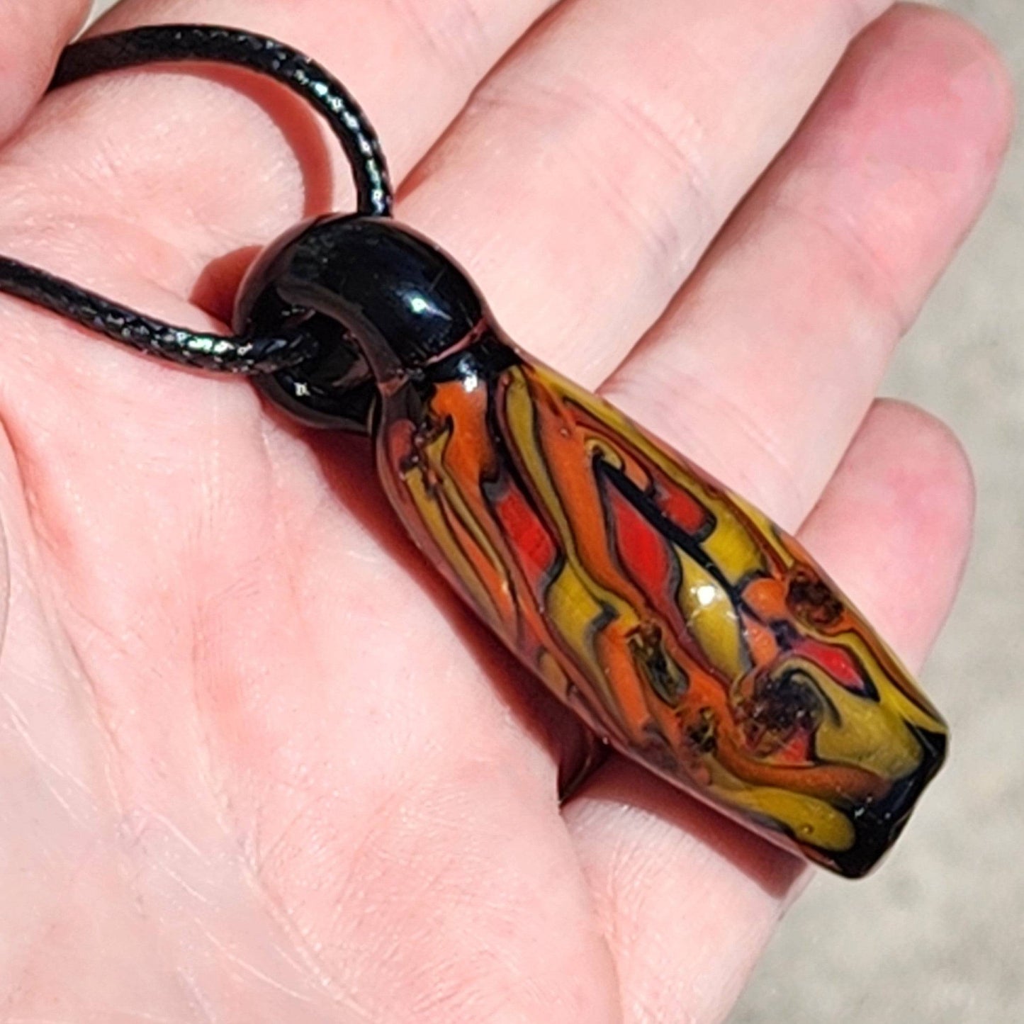 Handblown Tubular Glass Pendant with fiery swirl design held in hand, showing scale and detail. Handmade jewelry with black cord.
