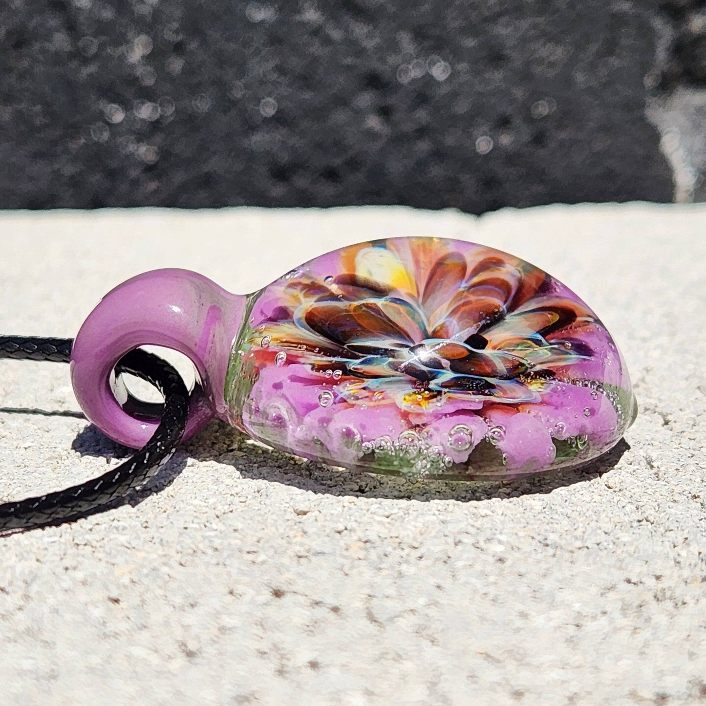 Low angle of handmade glass pendant with purple and orange flower design. Borosilicate necklace with black cord for jewelry collections.