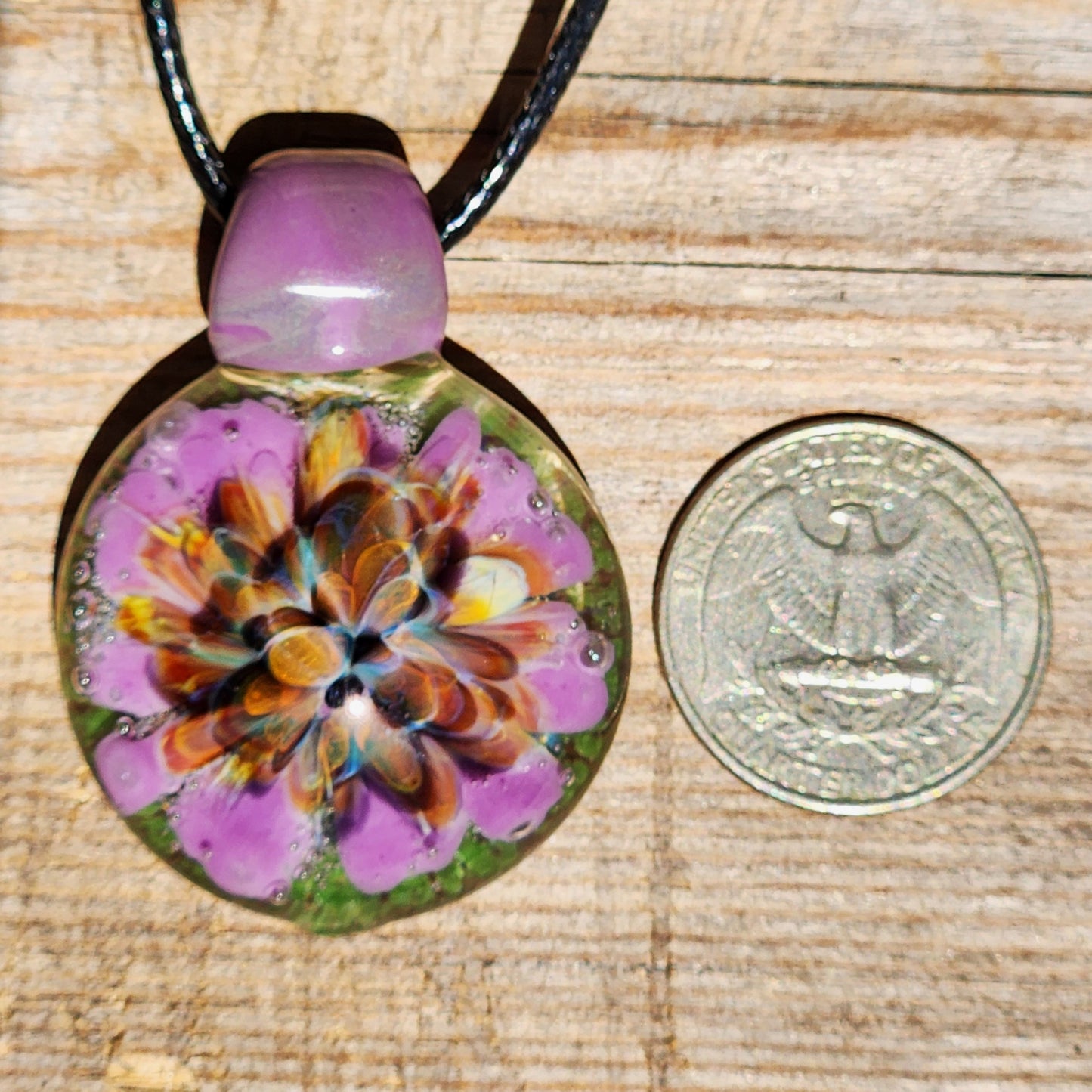 Size comparison of handmade glass pendant next to US quarter. Multicolor borosilicate jewelry with purple flower design.