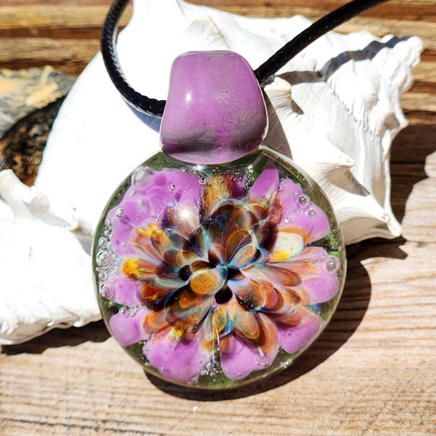 Handmade multicolor glass pendant resting on shell, showcasing intricate flower design. Borosilicate jewelry for unique necklace lovers.