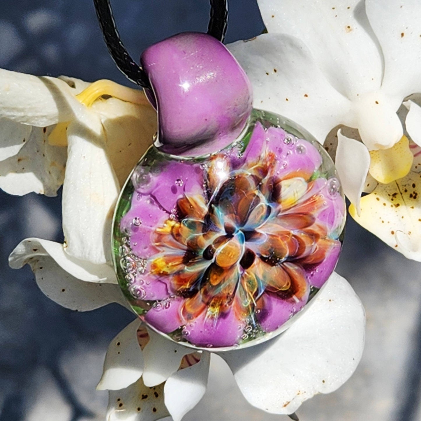 Close-up of handmade borosilicate pendant hanging on flower, showcasing vibrant purple and orange floral design. Heady glass necklace.