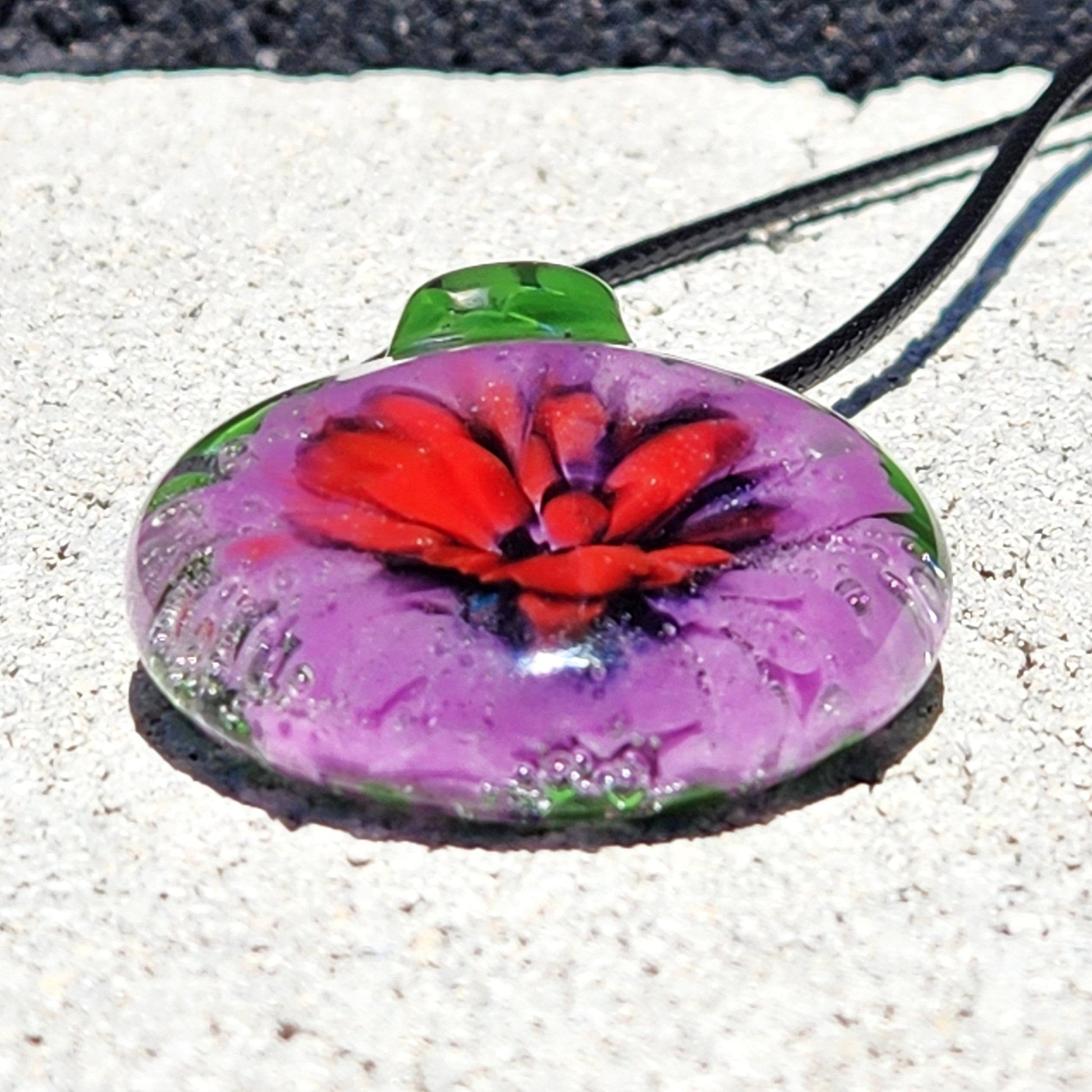 Large Heady Glass Pendant Necklace lying flat, showcasing red and purple flower in green glass. Handmade blown glass jewelry with artistic flair.