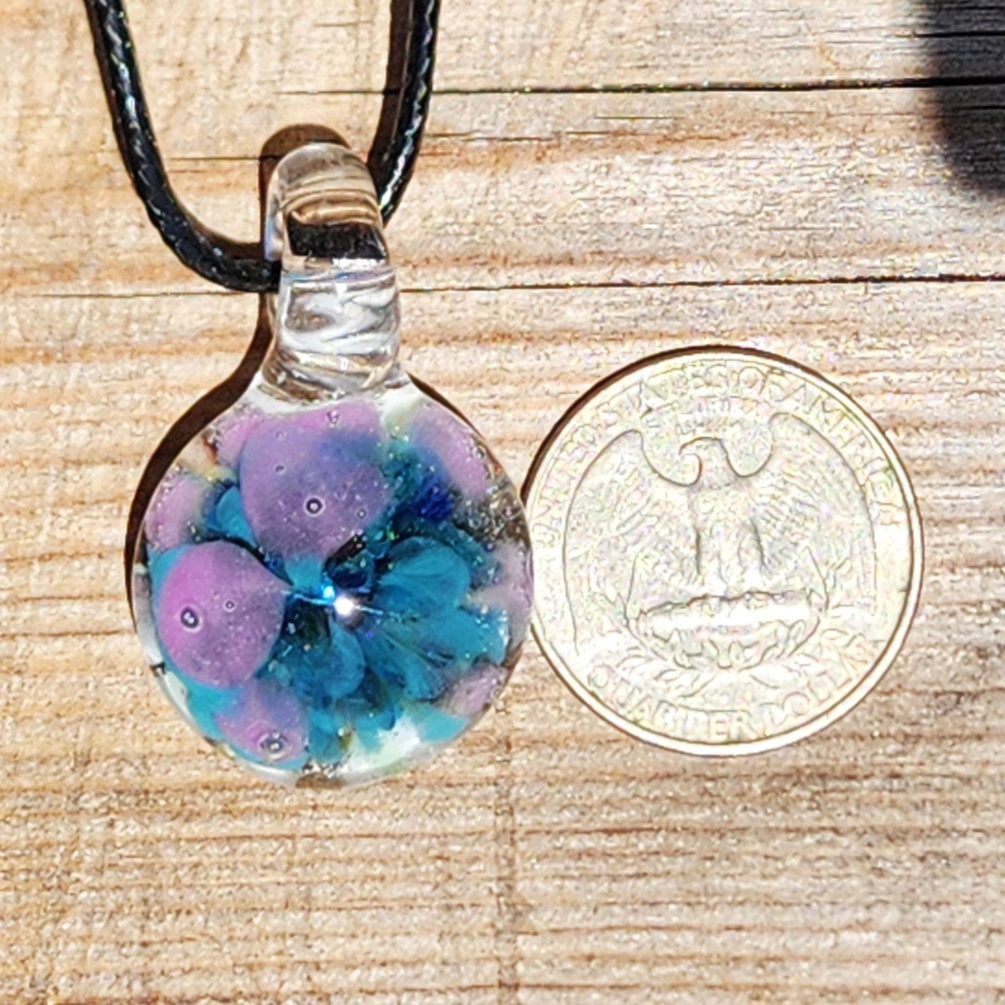 Small handmade glass pendant next to US quarter for size comparison. Features purple and blue floral design in borosilicate glass.