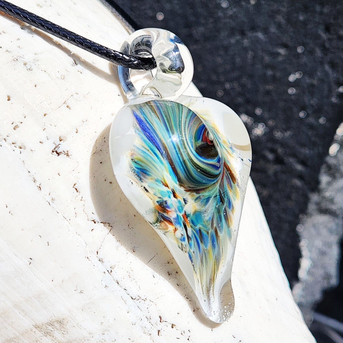 Unique trippy heart pendant made from blown borosilicate glass with colorful swirling patterns, shown from a side angle on a shell.